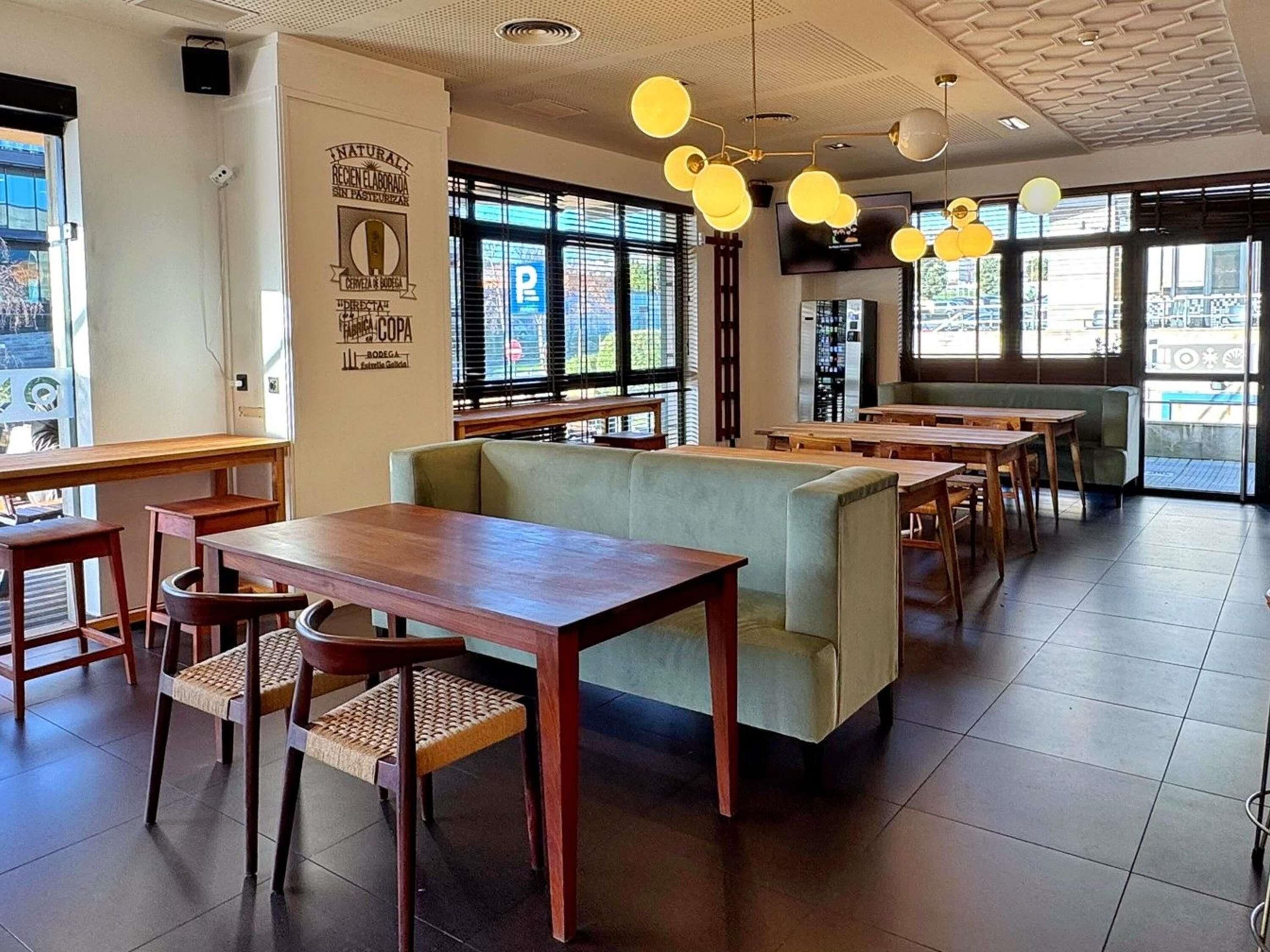 A restaurant interior with wooden tables and chairs, a green cushioned bench, and large windows with a view of the street.