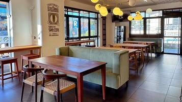 A restaurant interior with wooden tables and chairs, a green cushioned bench, and large windows with a view of the street.