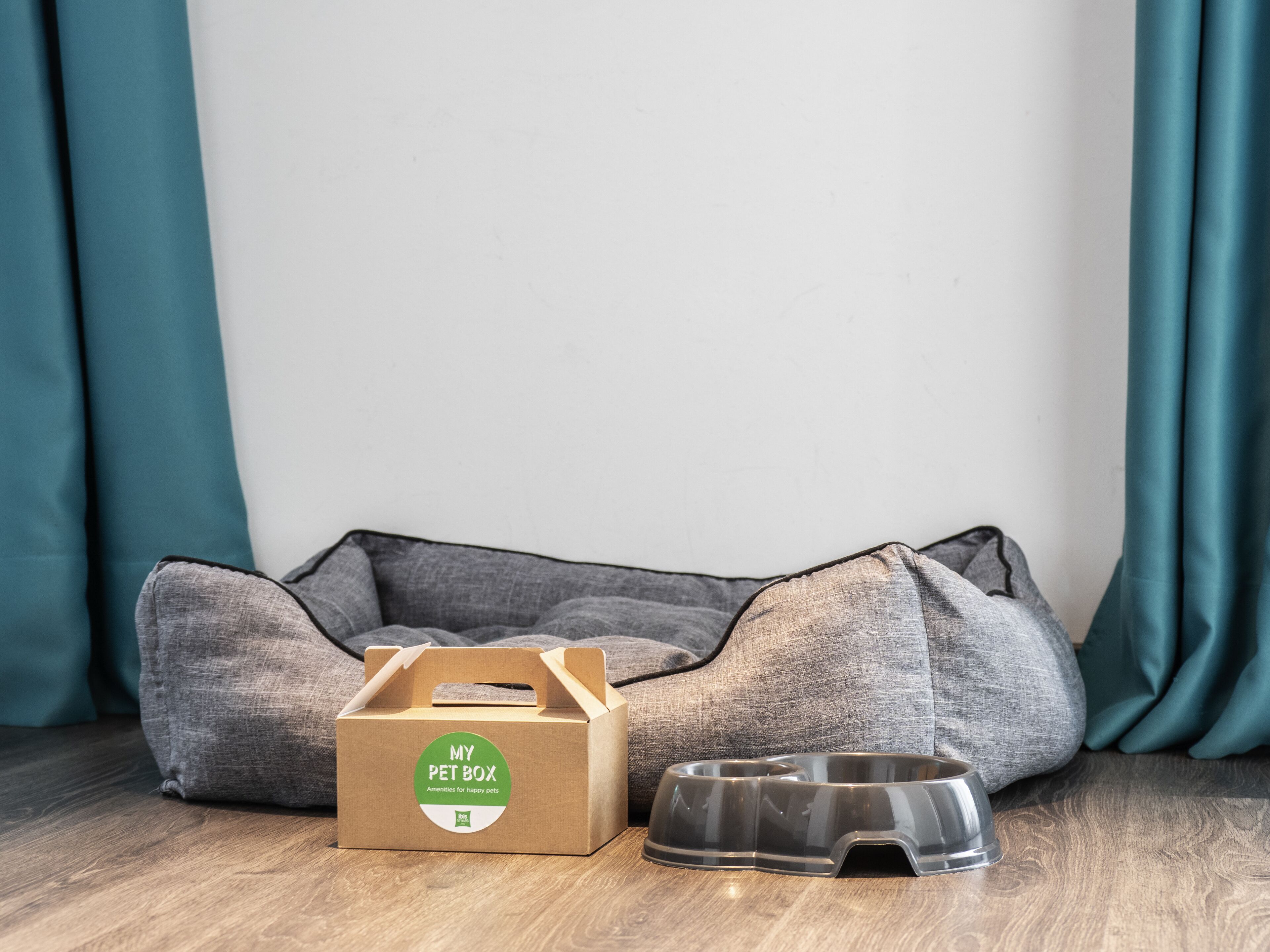 Una cama para mascotas, una caja de cartón con la etiqueta "MY PET BOX" y un cuenco de comida para mascotas sobre un piso de madera.