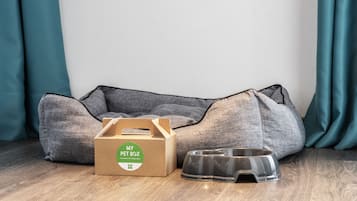 Una cama para mascotas, una caja de cartón con la etiqueta "MY PET BOX" y un cuenco de comida para mascotas sobre un piso de madera.
