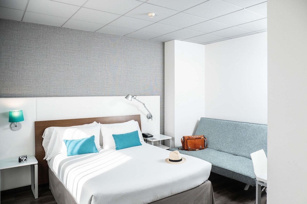 Ein modernes Hotelzimmer mit einem aufgeräumten Bett, einer Nachttischlampe, einem kleinen Tisch, einem Sessel und einem Sofa.