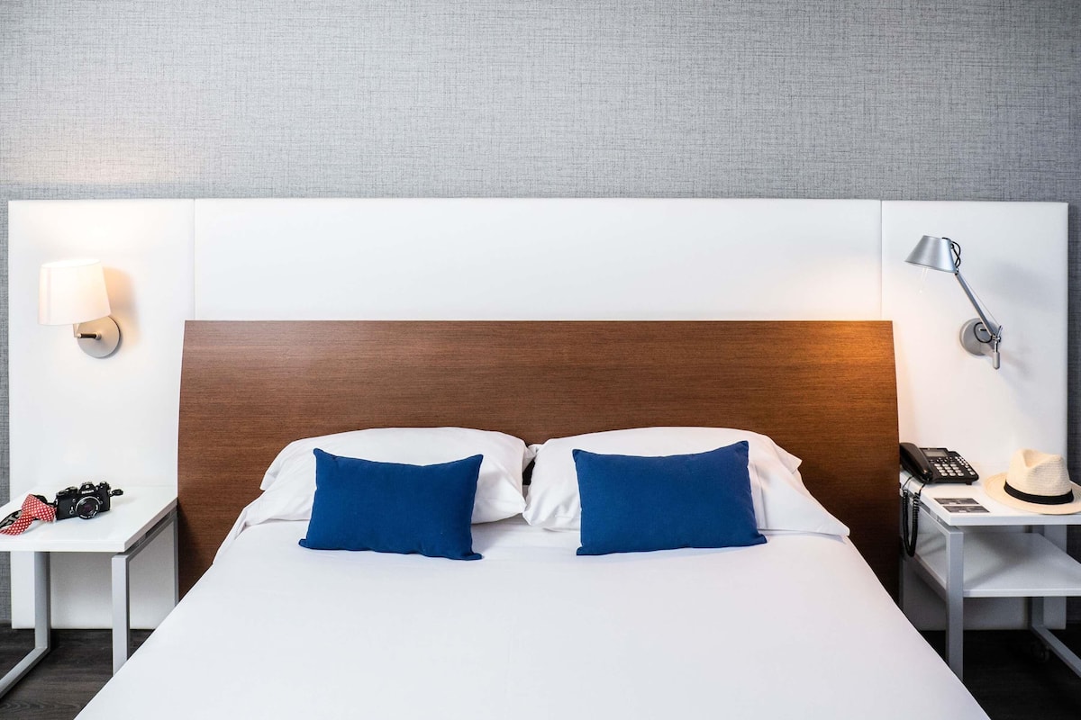 Ein Hotelzimmer mit einem großen Bett, zwei Kissen, einem Holz-Kopfteil, einem Nachttisch mit Lampe, einem Telefon und einer Kamera auf dem Beistelltisch.