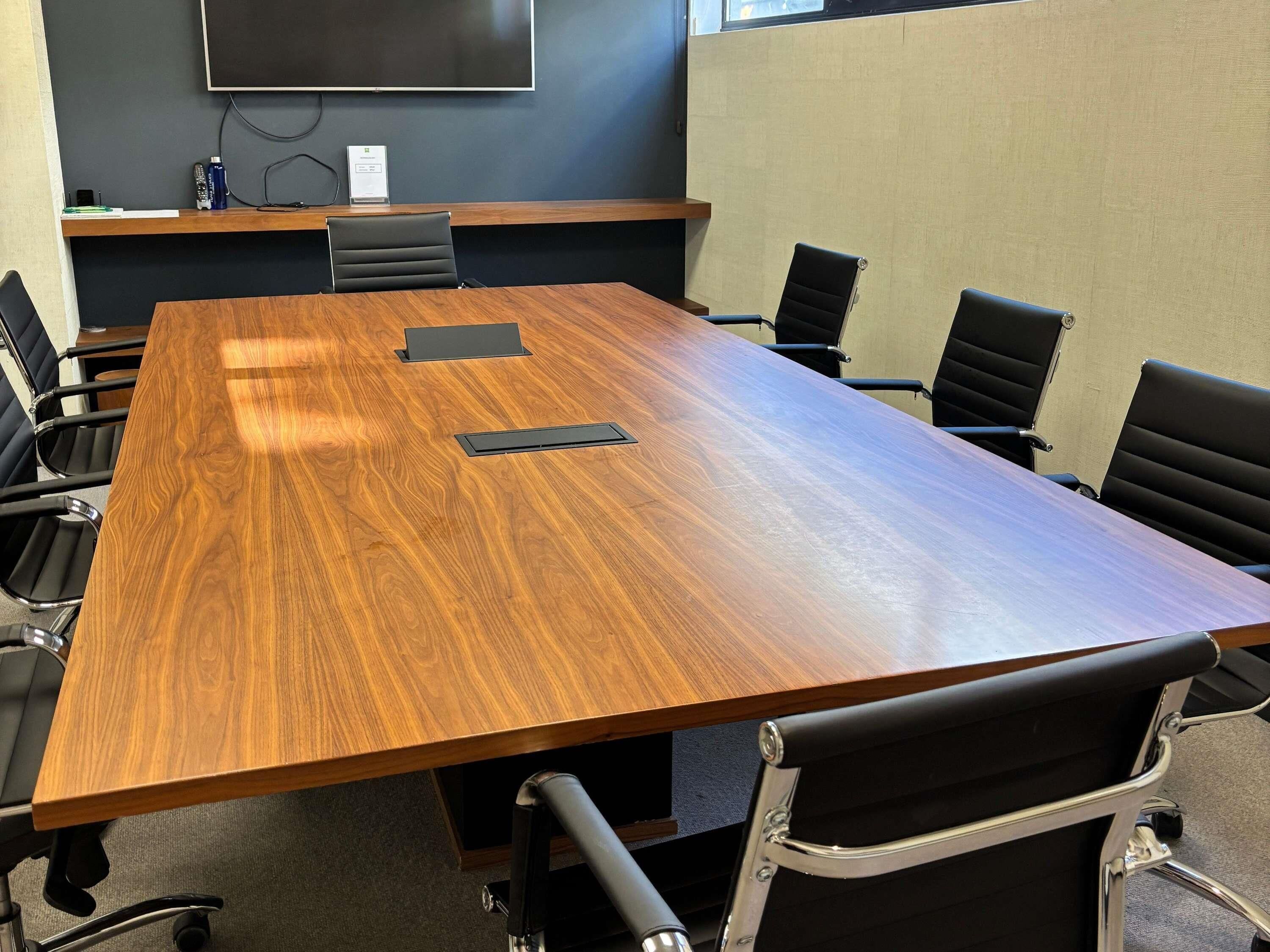 Un salón de conferencias con una gran mesa de madera, sillas negras, una pantalla para proyector y un estante con objetos.