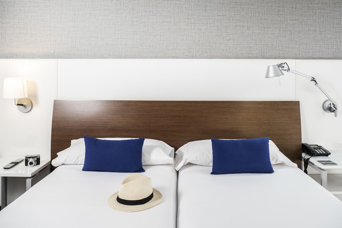 Ein Hotelzimmer mit einem hölzernen Kopfteil, zwei blauen Kissen, einer weißen Bettdecke, einem Strohhut, einer Nachttischlampe, einem Telefon und einer Fernbedienung auf dem Nachttisch.