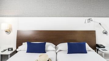 A hotel room with a wooden headboard, two blue pillows, a white bedspread, a straw hat, a bedside lamp, a phone, and a remote control on the nightstand.