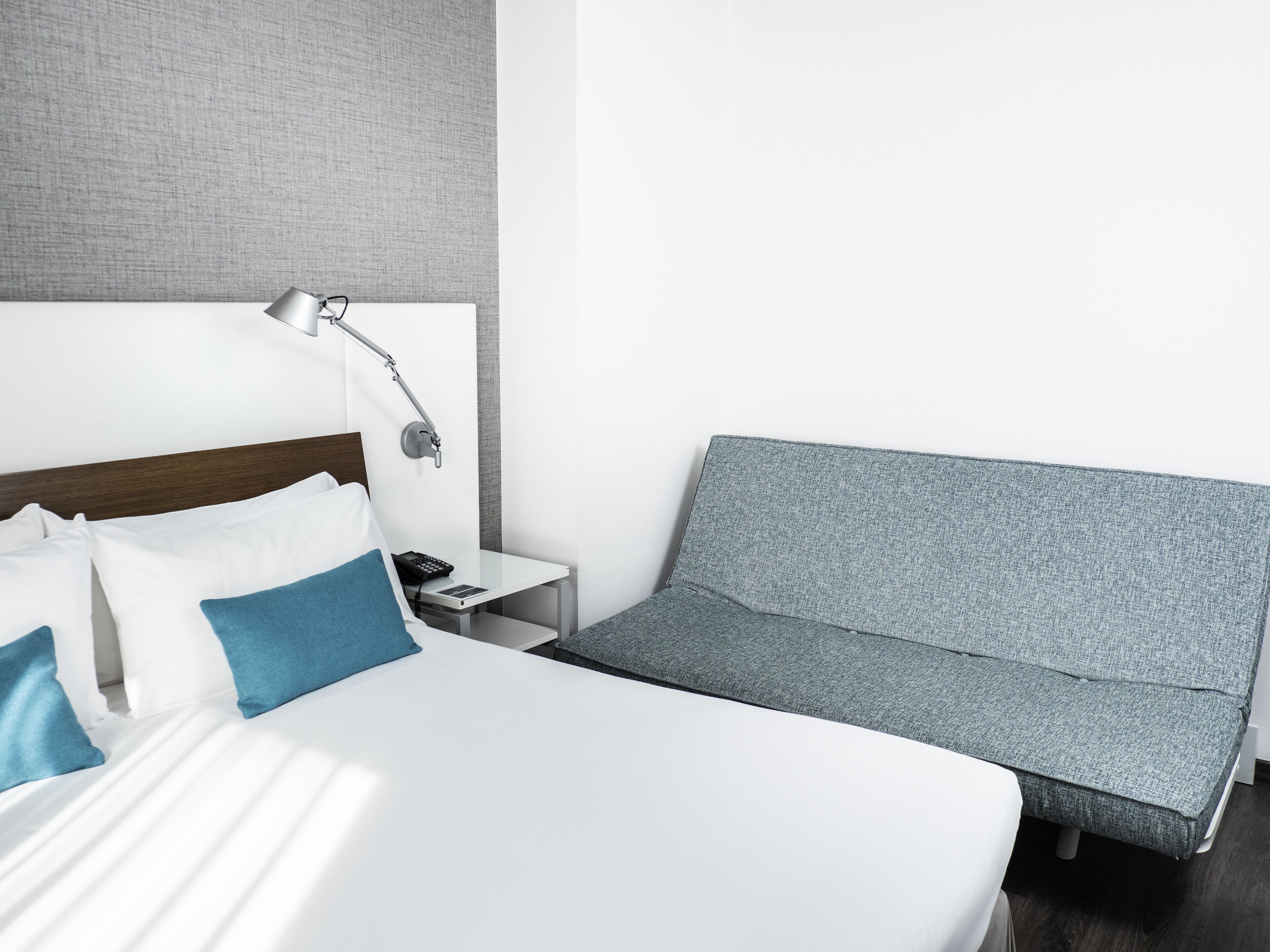 Una habitación de hotel moderna con una cama bien hecha, un sofá gris, una mesita de noche con lámpara y un teléfono instalado en la pared.