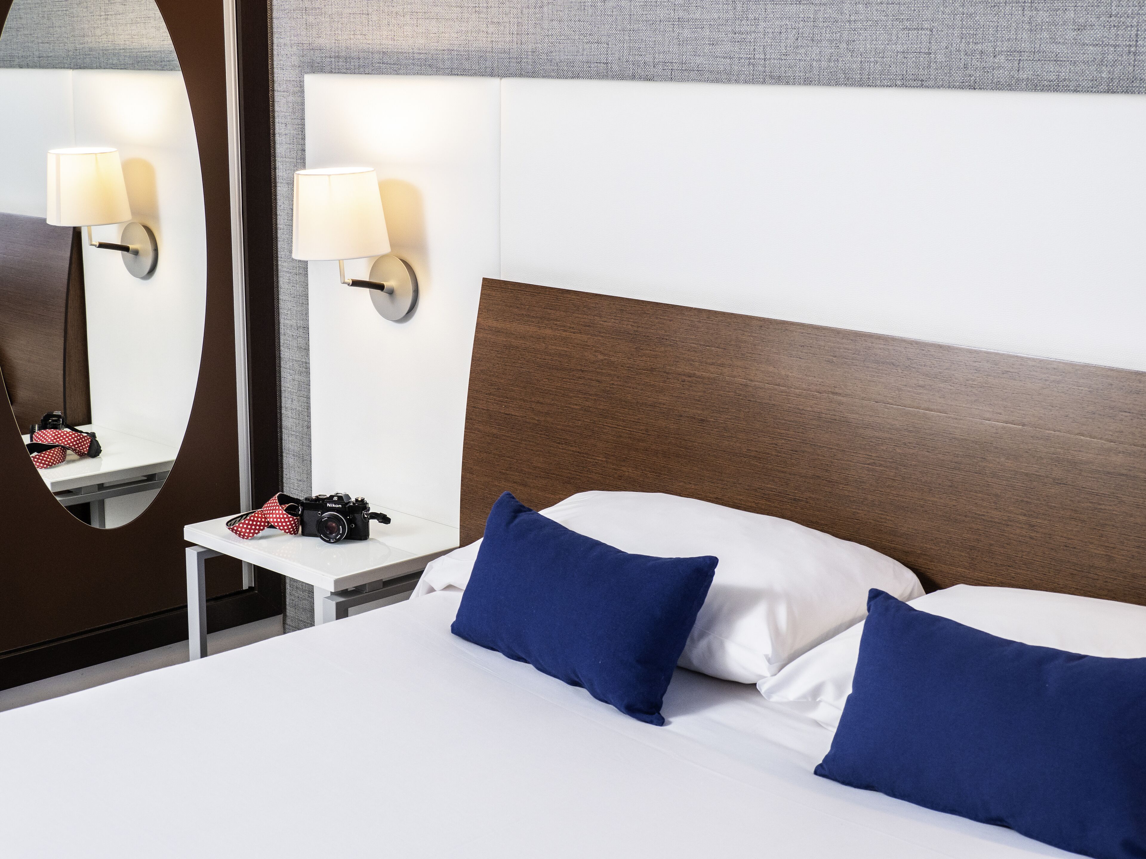 A hotel room with a wooden headboard, two pillows, a nightstand with a lamp, and a mirror.
