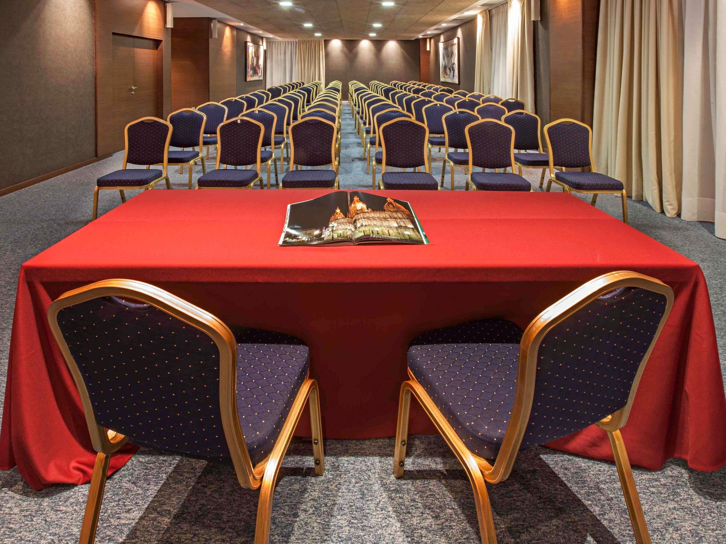 Un salón de conferencias con filas de sillas, una mesa roja y un folleto.