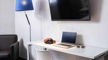 Une chambre moderne avec un téléviseur à écran plat fixé au mur, un bureau avec un ordinateur portable, un appareil photo et des lunettes de soleil, ainsi qu’une lampe de sol bleue.