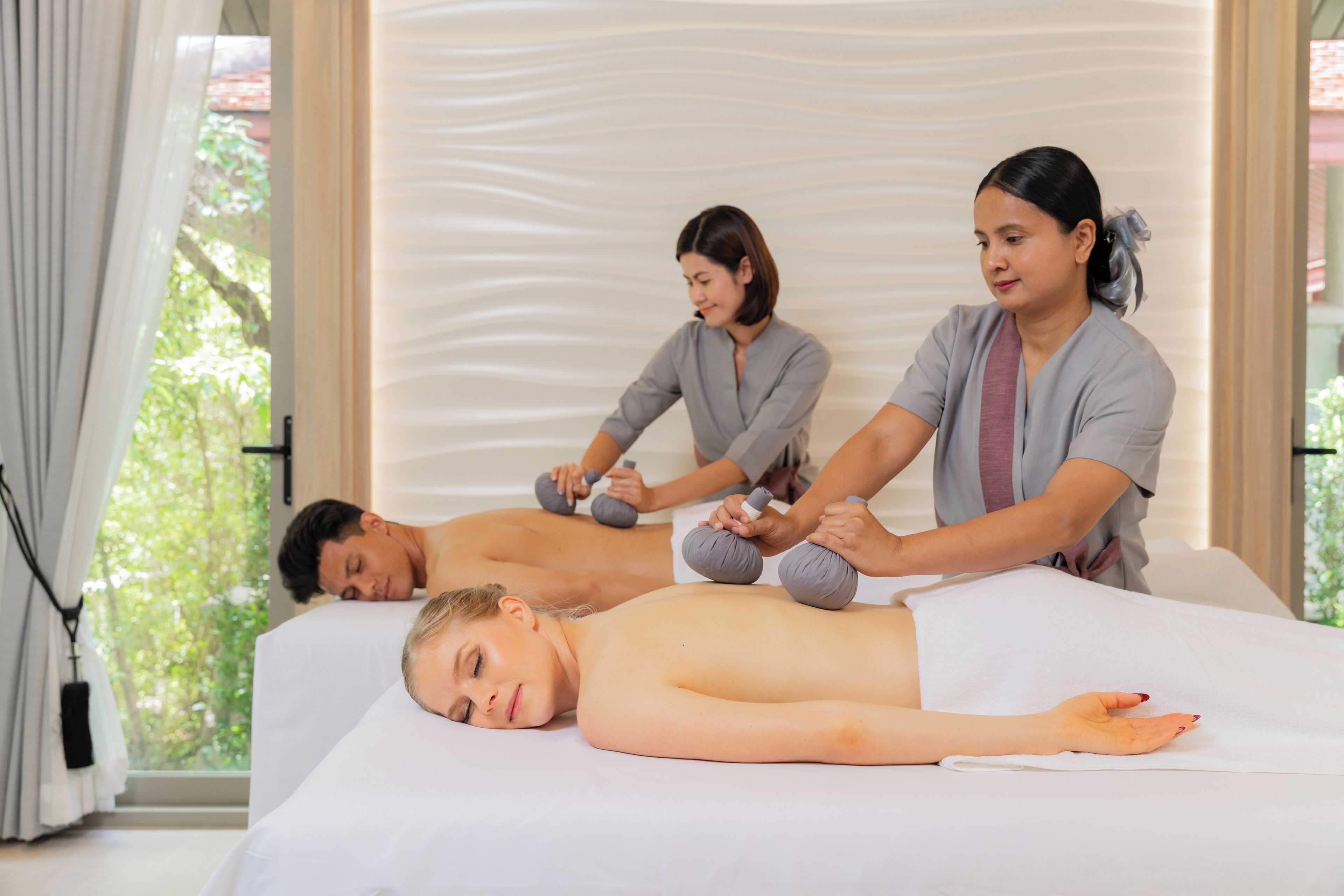 Eine Massageanwendung in einem Raum mit großem Fenster, einem Massagetisch und einem Masseur, der mit einem Gerät Druck ausübt.