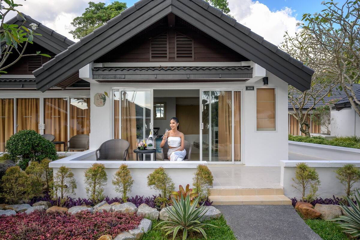 Une maison moderne dotée d’un balcon, d’un coin repas avec une table et des chaises, et d’un jardin agrémenté de diverses plantes et de rochers.