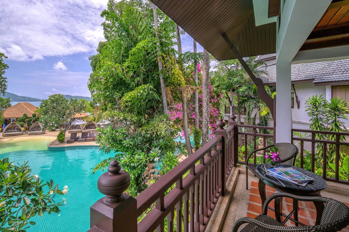 Un balcon donnant sur une piscine, une végétation luxuriante et une maison traditionnelle en arrière-plan.