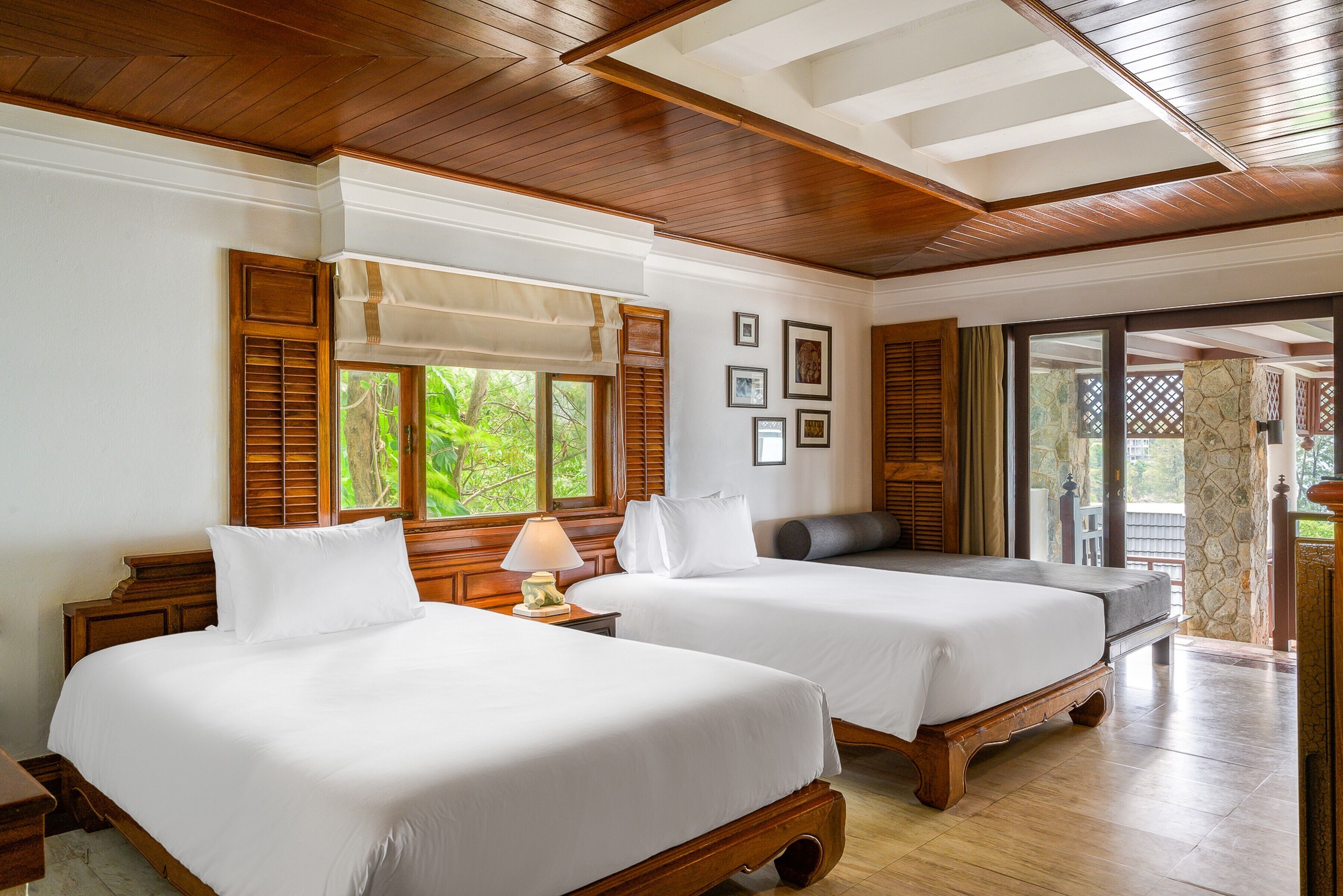 Une chambre d’hôtel avec deux lits, des têtes de lit en bois, une grande fenêtre avec volets, un mur en pierre et une vue sur la verdure environnante.