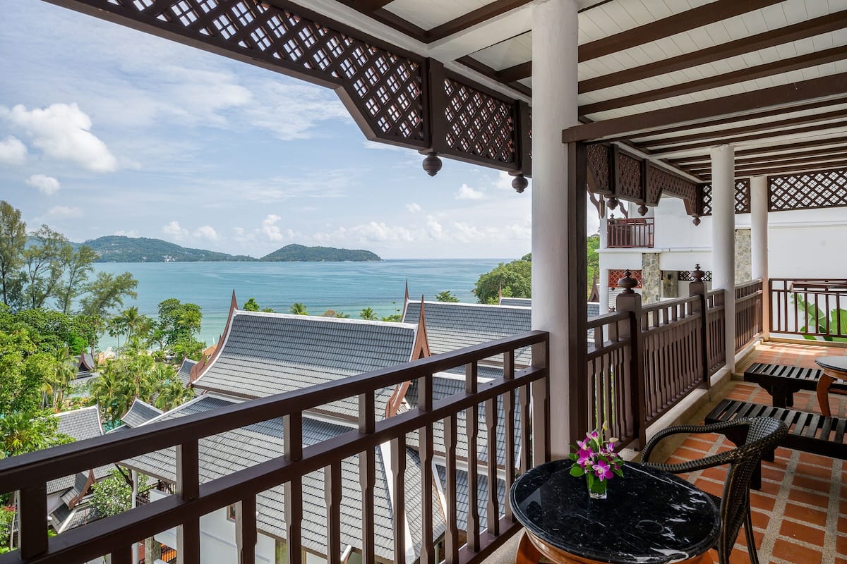 Un balcon avec vue sur la mer et les collines, des balustrades en bois et une table agrémentée d’un vase de fleurs.