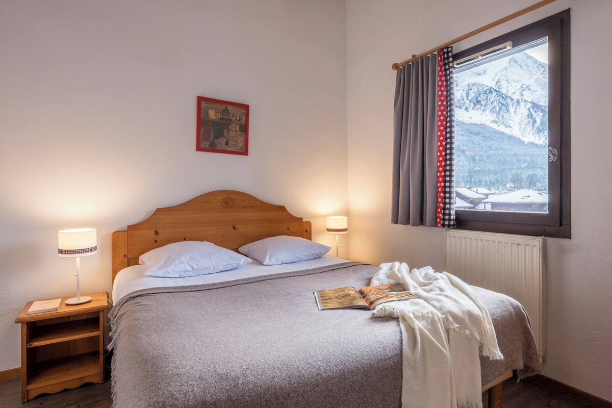A bedroom with a wooden headboard, two bedside lamps, a window with curtains, a radiator, and a bed with a blanket and a book.