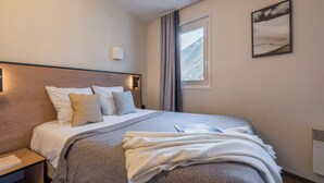 A neatly made bed with a grey blanket and pillows, a bedside table with a book, a wall-mounted picture, and a window with curtains. - Résidence Pierre & Vacances Chamonix La Rivière (Chamonix-Mont-Blanc)