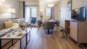 A modern living room with a sofa, coffee table, dining table with chairs, a TV, and a balcony view. - Résidence Pierre & Vacances Chamonix La Rivière (Chamonix-Mont-Blanc)