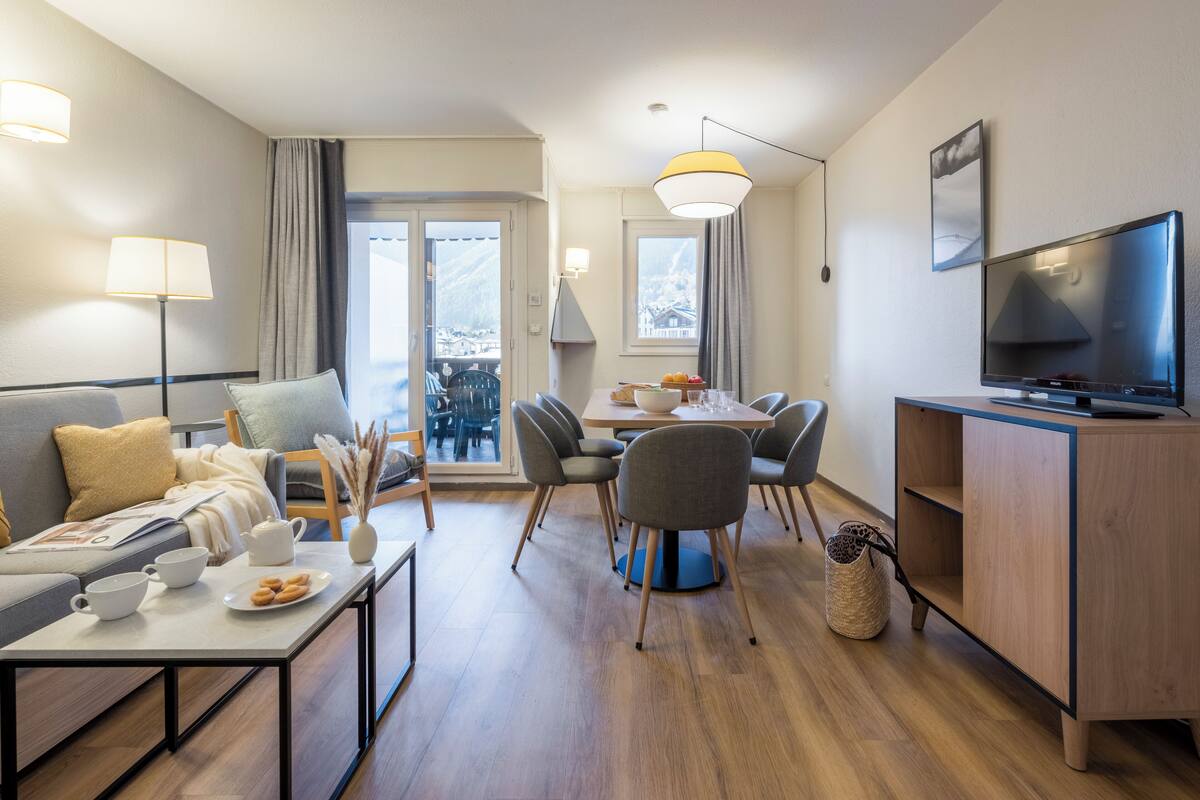 A modern living room with a sofa, coffee table, dining table with chairs, a TV, and a balcony view.