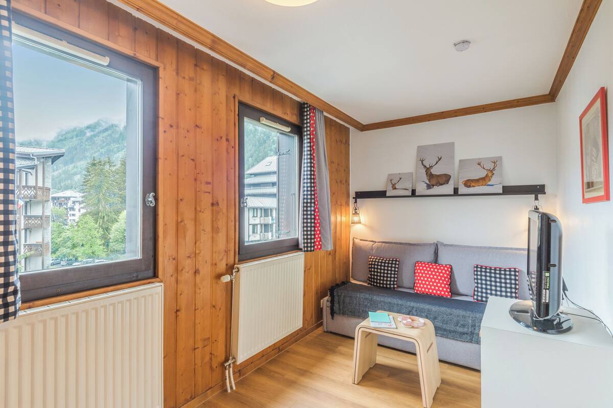 A cozy room with wooden walls, a grey sofa with patterned cushions, a small table, and a mounted TV.