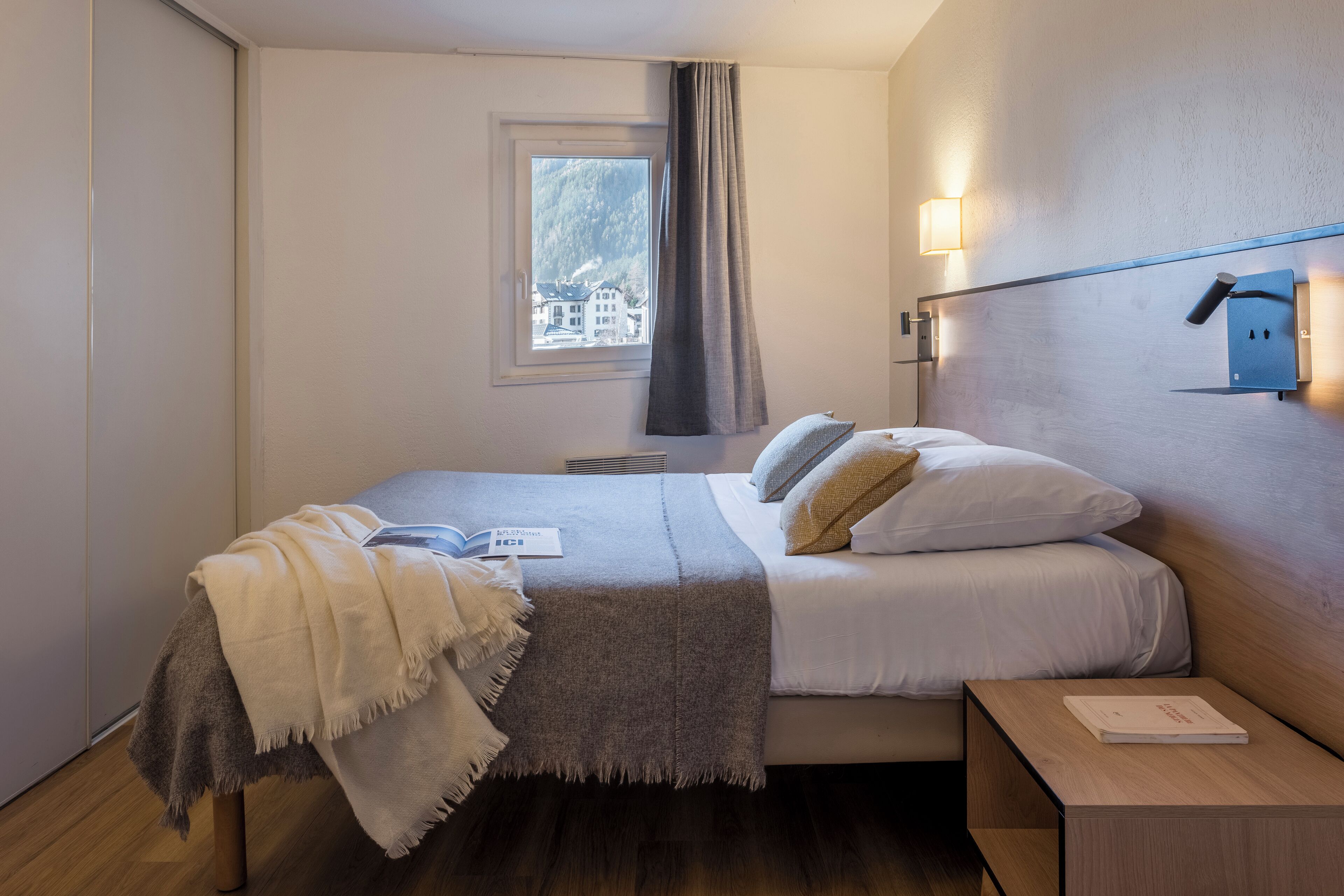 A hotel room with a bed, a bedside table, a window with a view of buildings, and a wall-mounted light fixture.