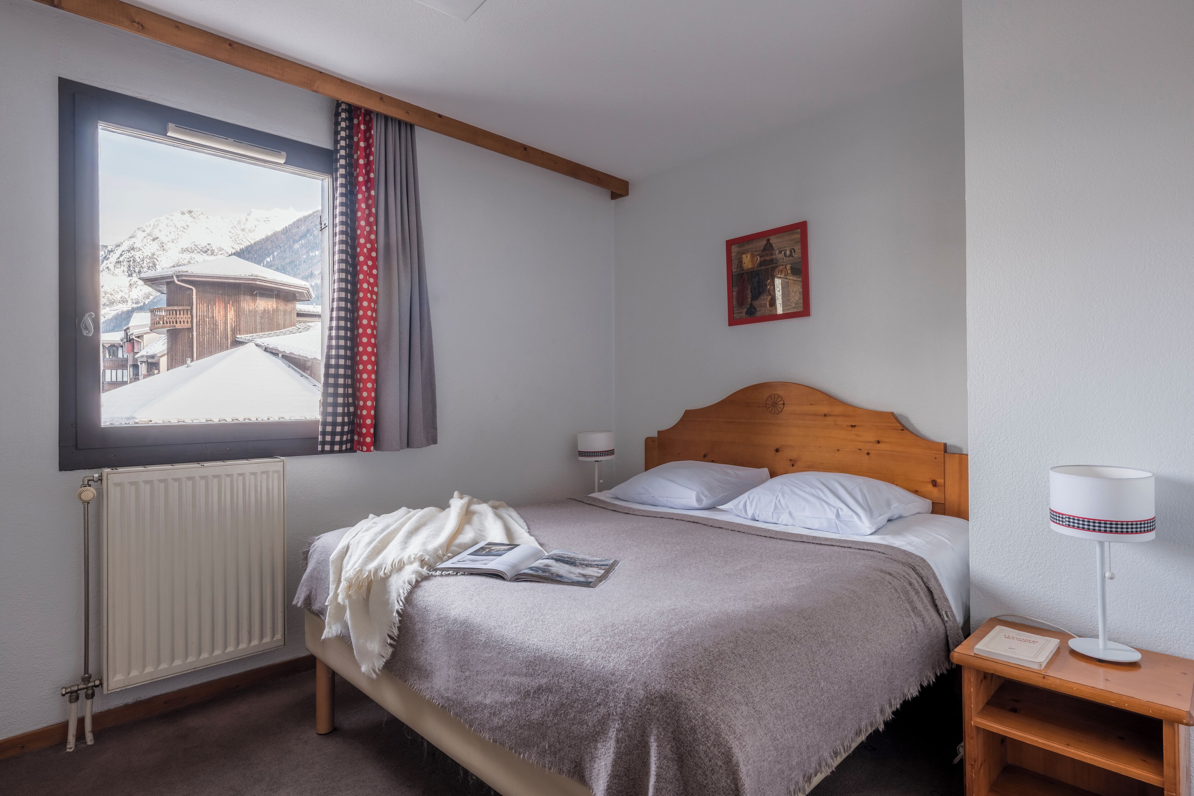 Une chambre à coucher avec un lit, une fenêtre donnant sur des bâtiments enneigés, un radiateur, une table de chevet avec une lampe et un tableau encadré accroché au mur.