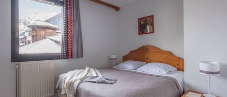 Une chambre à coucher avec un lit, une fenêtre donnant sur des bâtiments enneigés, un radiateur, une table de chevet avec une lampe et un tableau encadré accroché au mur.