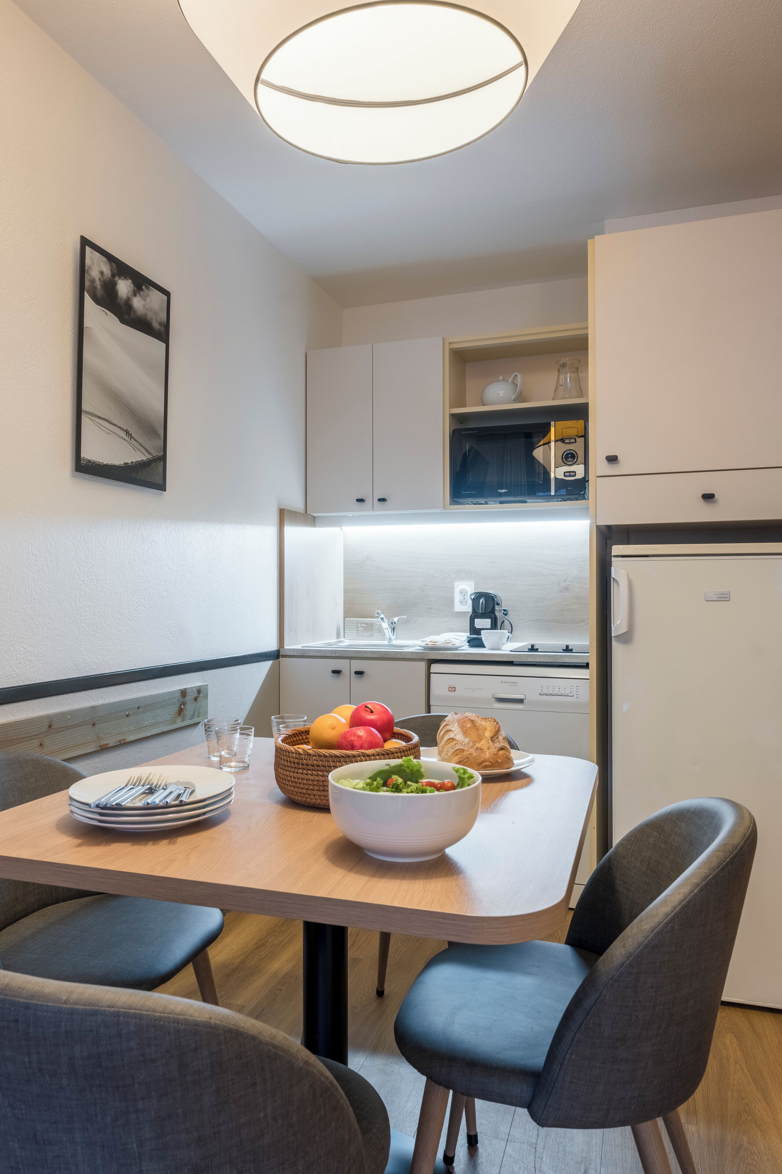 Een moderne keuken met witte kastjes, een inbouwoven en een koelkast. Er staat een eettafel met stoelen, een schaal met fruit en een mand met groenten.