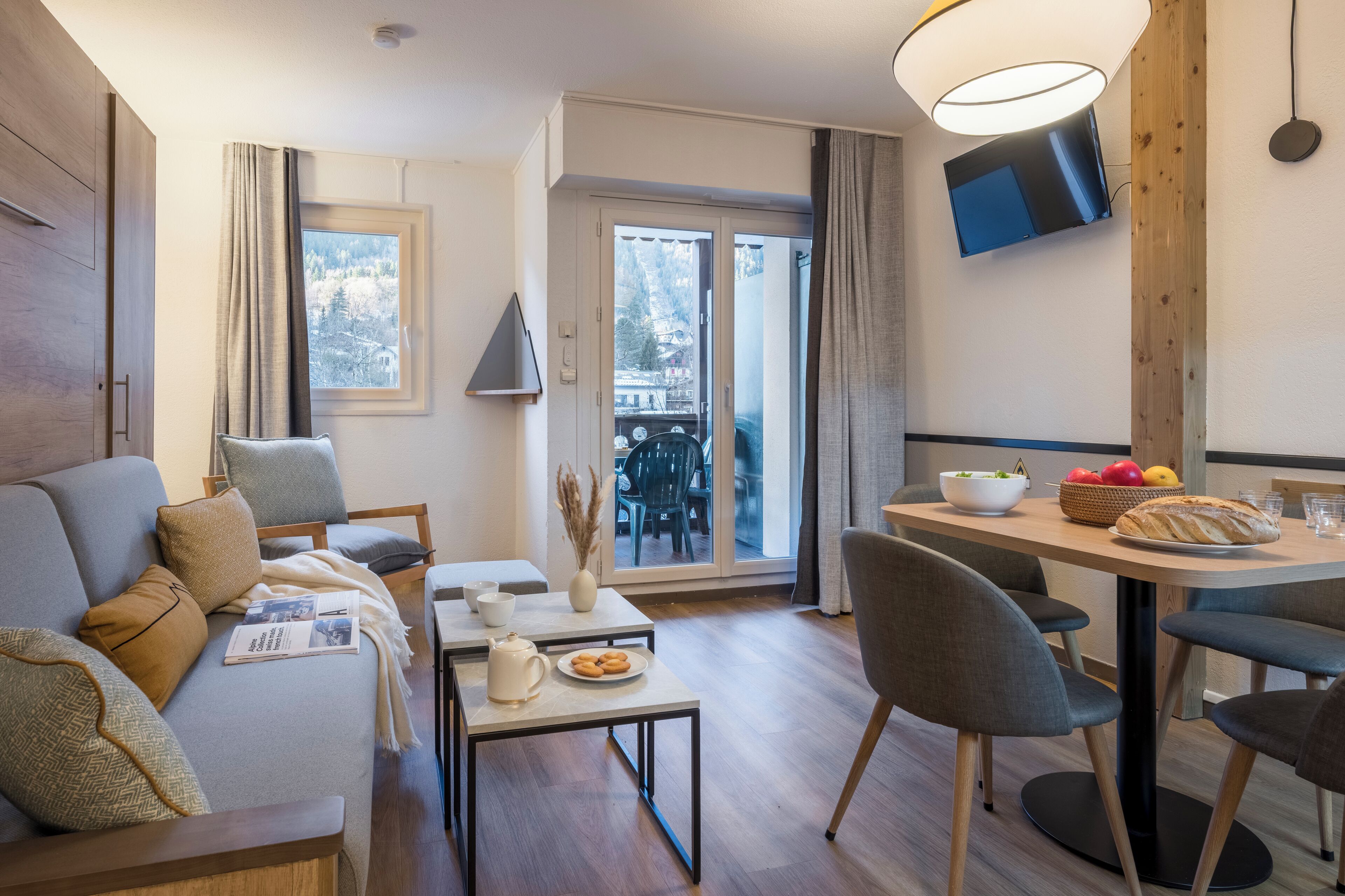 Een moderne woonkamer met een bank, een eettafel met stoelen, een flatscreen televisie aan de muur en uitzicht door de glazen deur naar buiten.