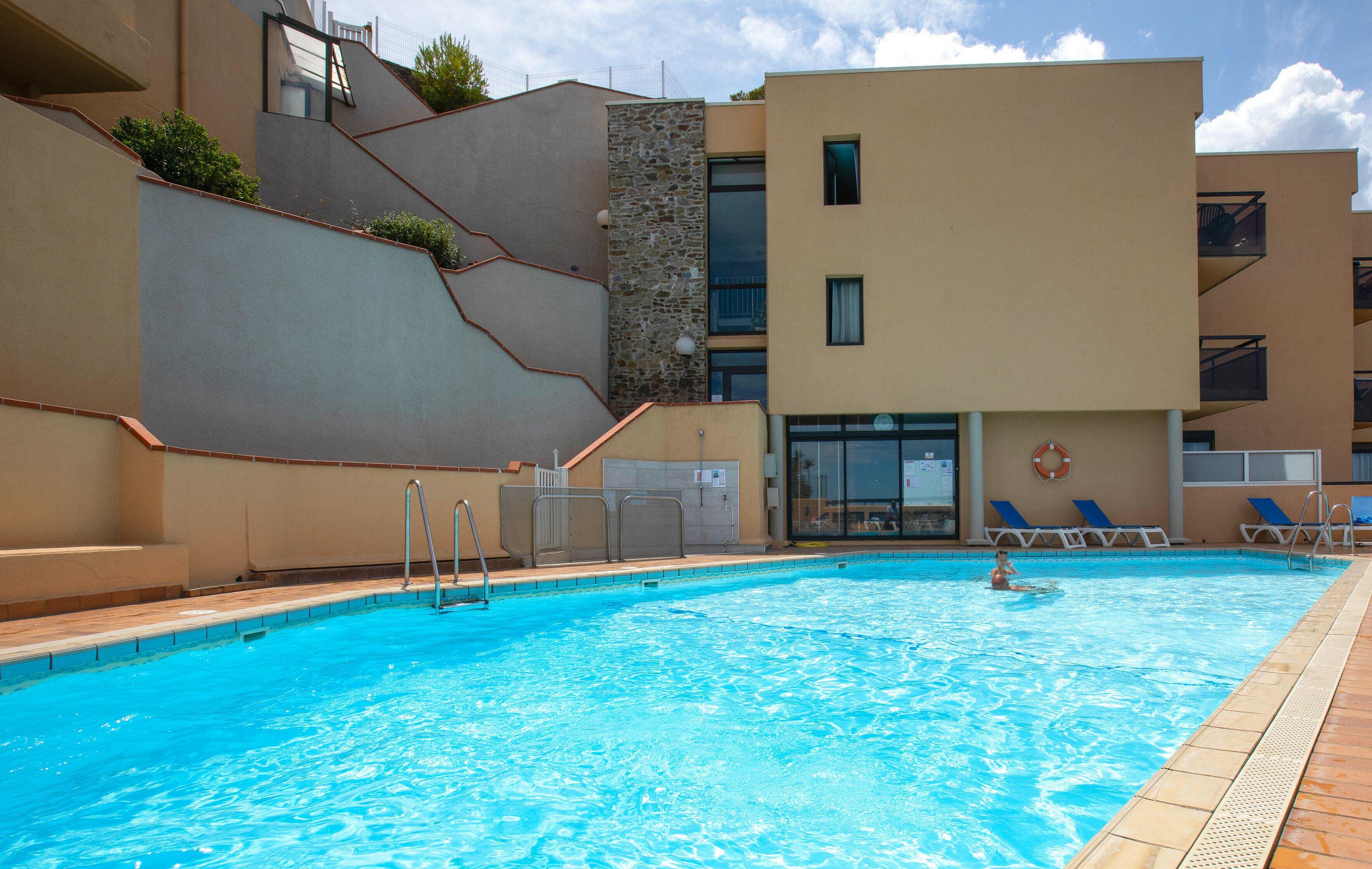 Une piscine à l’eau d’un bleu cristallin, entourée d’un bâtiment moderne doté de balcons et d’un poste de surveillance pour les sauveteurs.