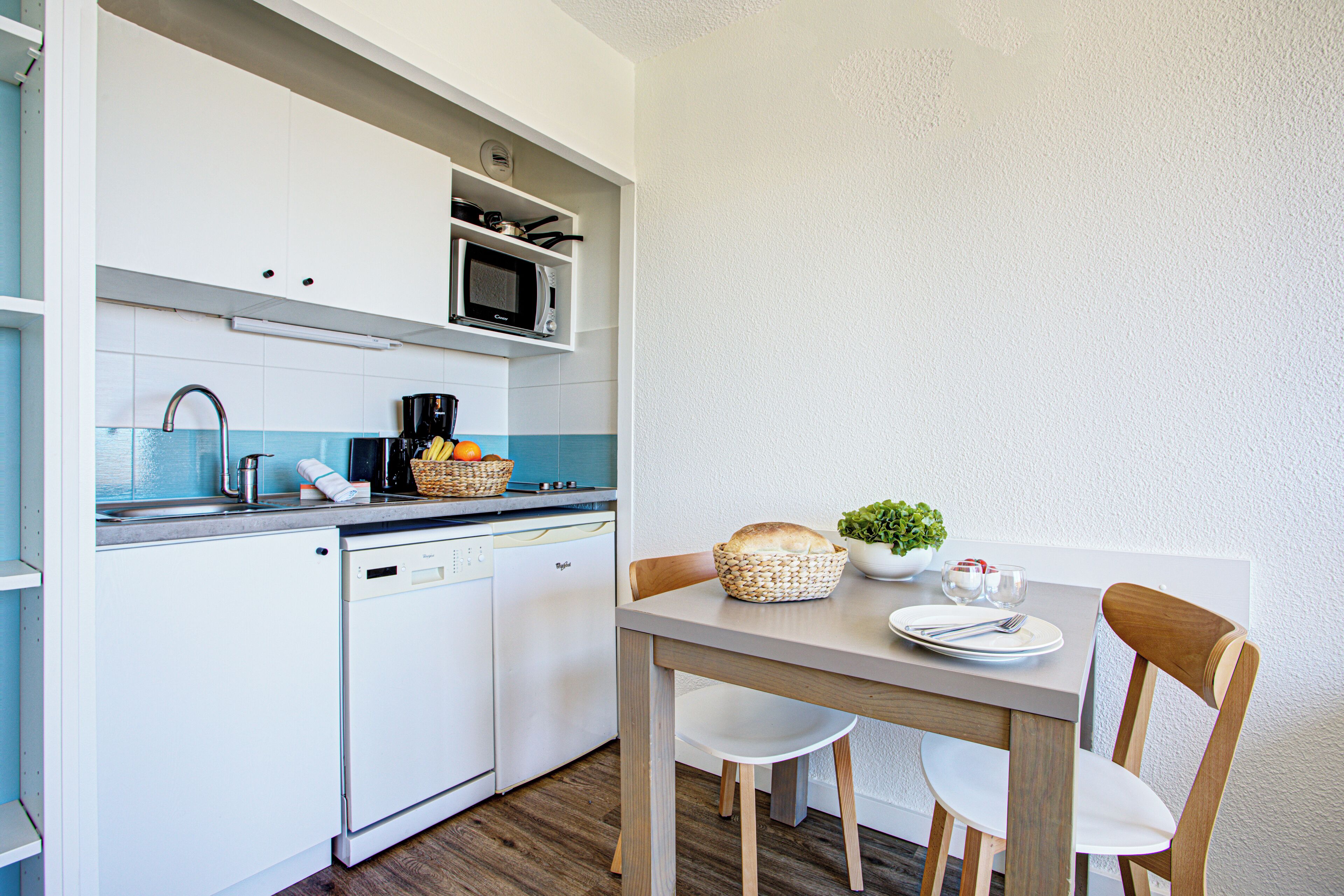 Une cuisine compacte avec des meubles blancs, un four à micro-ondes et un lave-vaisselle. On y trouve une table à manger avec des chaises et une petite plante posée sur la table.