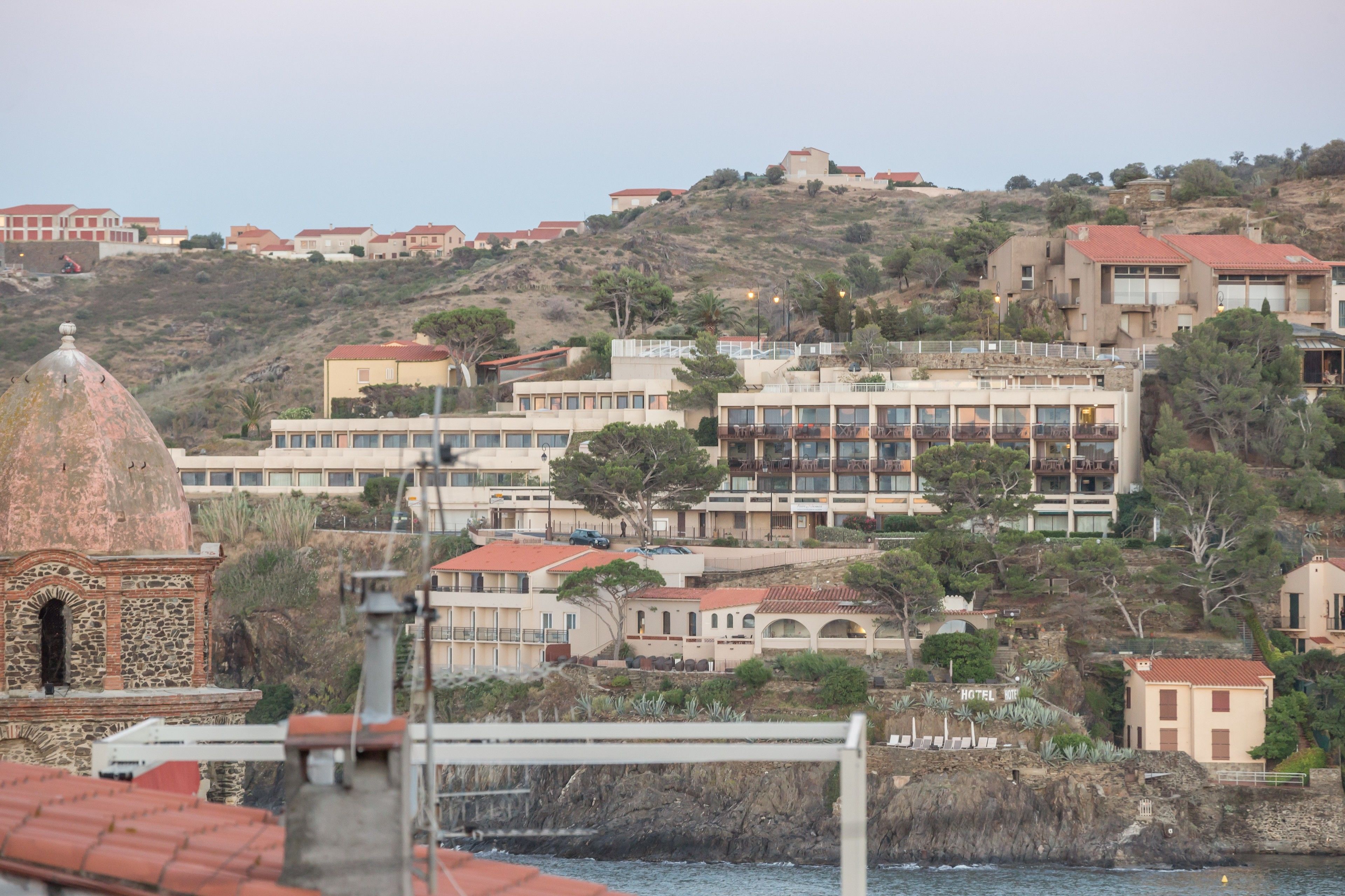 A coastal town with buildings on a hillside, including a hotel with balconies and a dome structure.