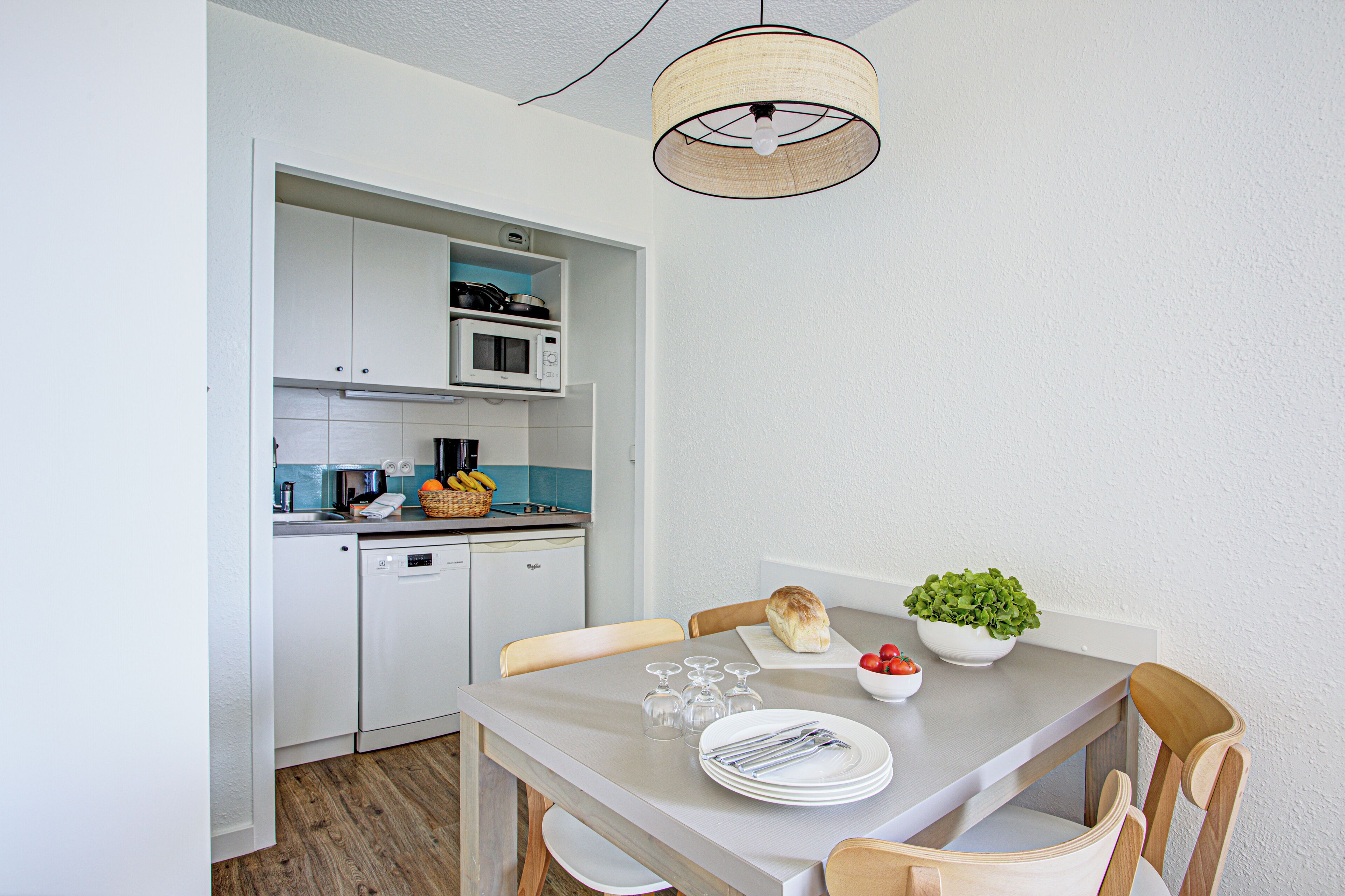 Une cuisine compacte avec des meubles blancs, un four à micro-ondes et un grille-pain. On y trouve une table à manger avec des chaises, un saladier rempli de fruits et une plante en pot.