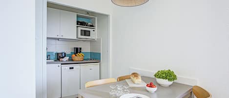 A compact kitchen with white cabinets, a microwave, and a toaster. There's a dining table with chairs, a bowl of fruit, and a potted plant.