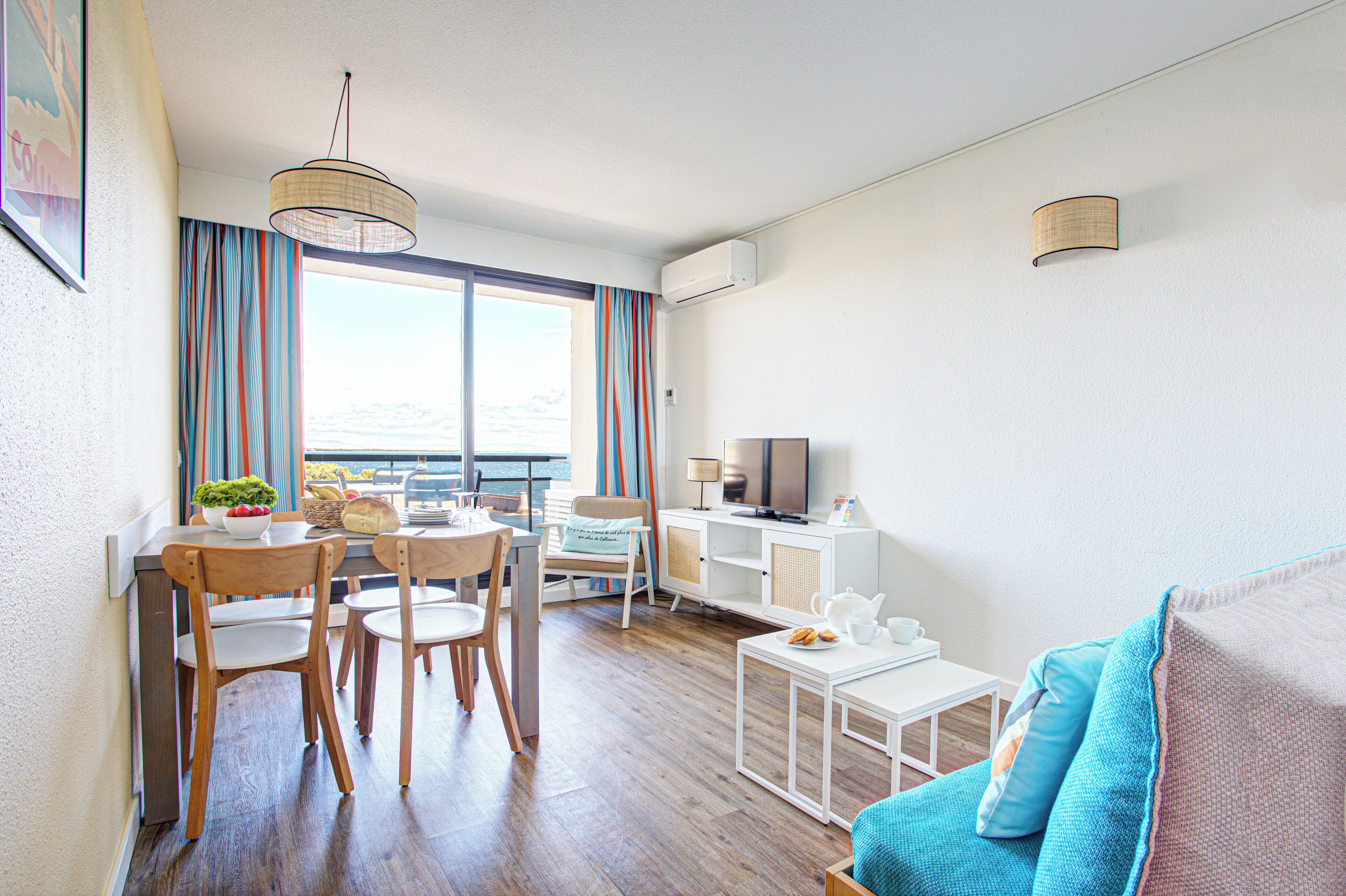 A modern living room with a dining area, a sofa, a TV, and a view of the sea.