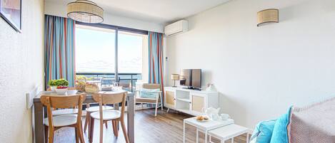 A modern living room with a dining area, a sofa, a TV, and a view of the sea.