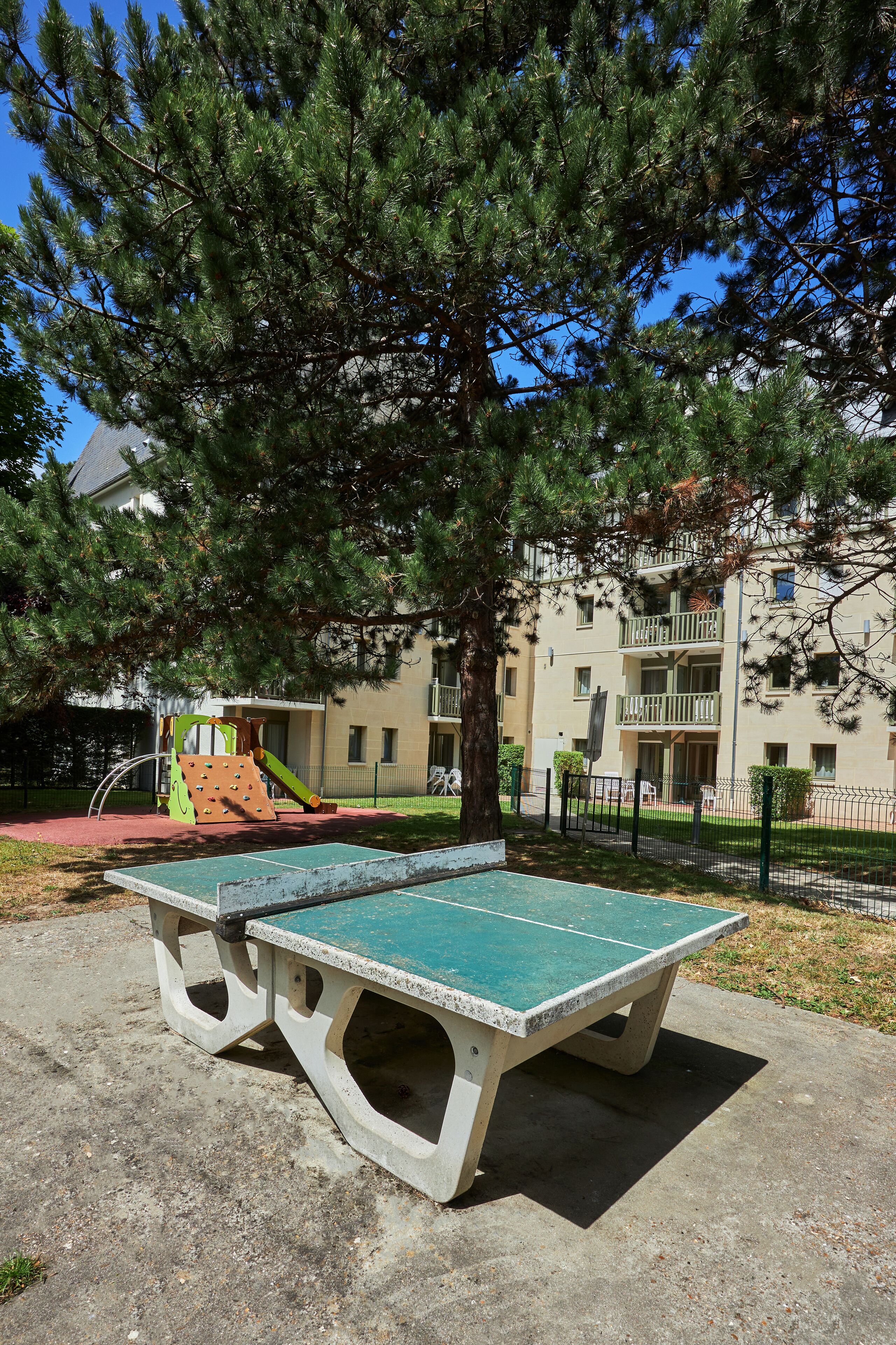 Una mesa de ping pong de hormigón en un área de parque con un parque infantil y edificios de apartamentos al fondo.