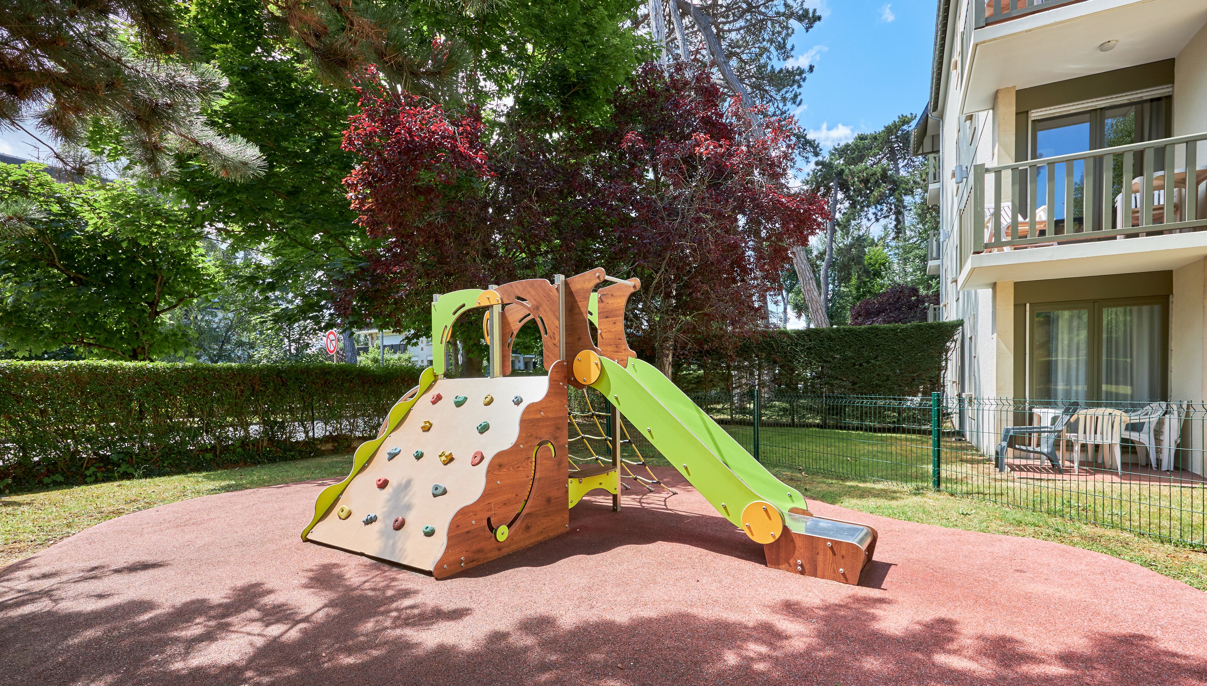 Un terrain de jeux équipé de structures d’escalade et de toboggans, entouré d’une haie et d’arbres.