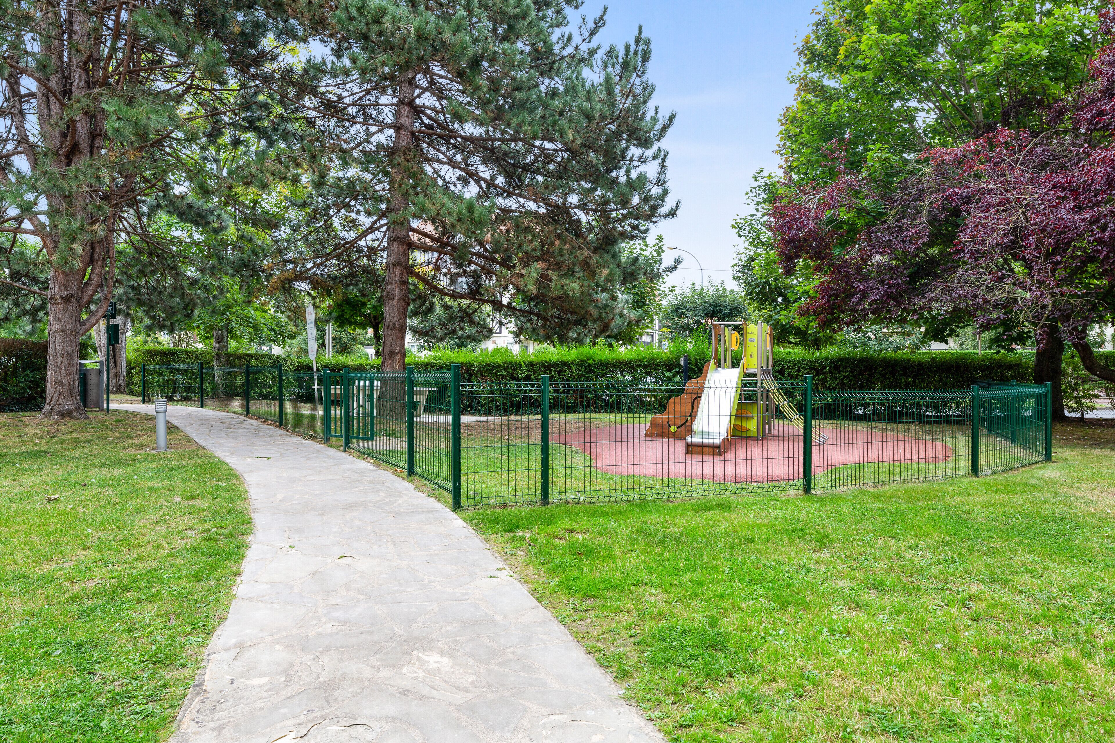 A park with a concrete pathway, green fence, and playground equipment.