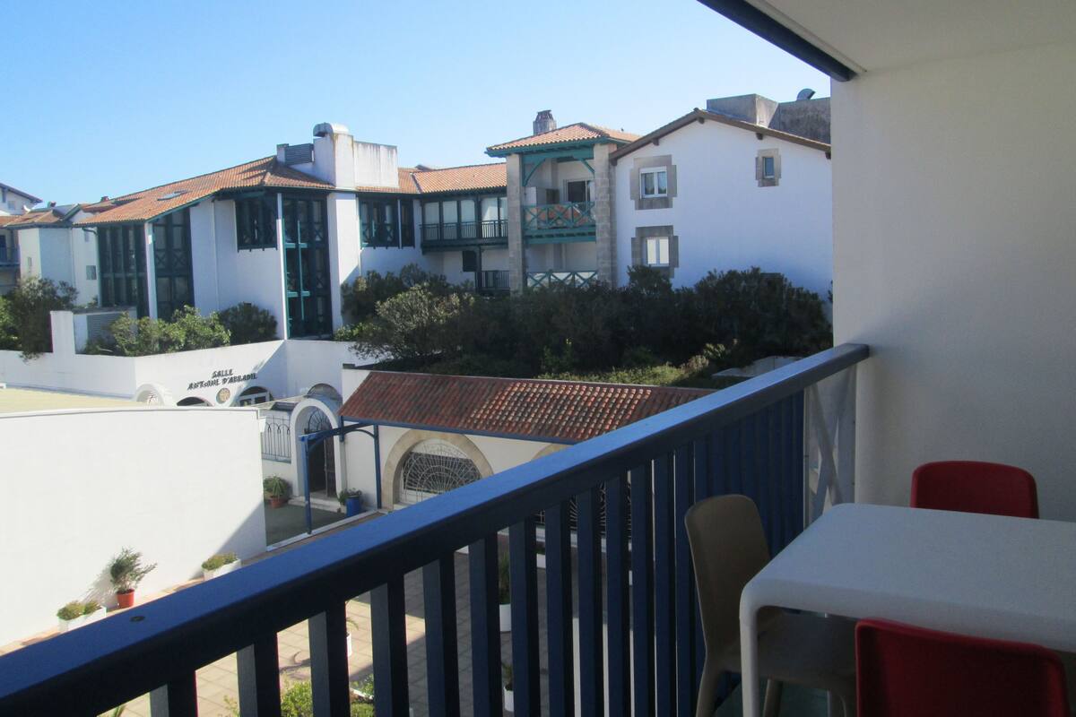 A balcony with a table and chairs, overlooking a courtyard with buildings and greenery.