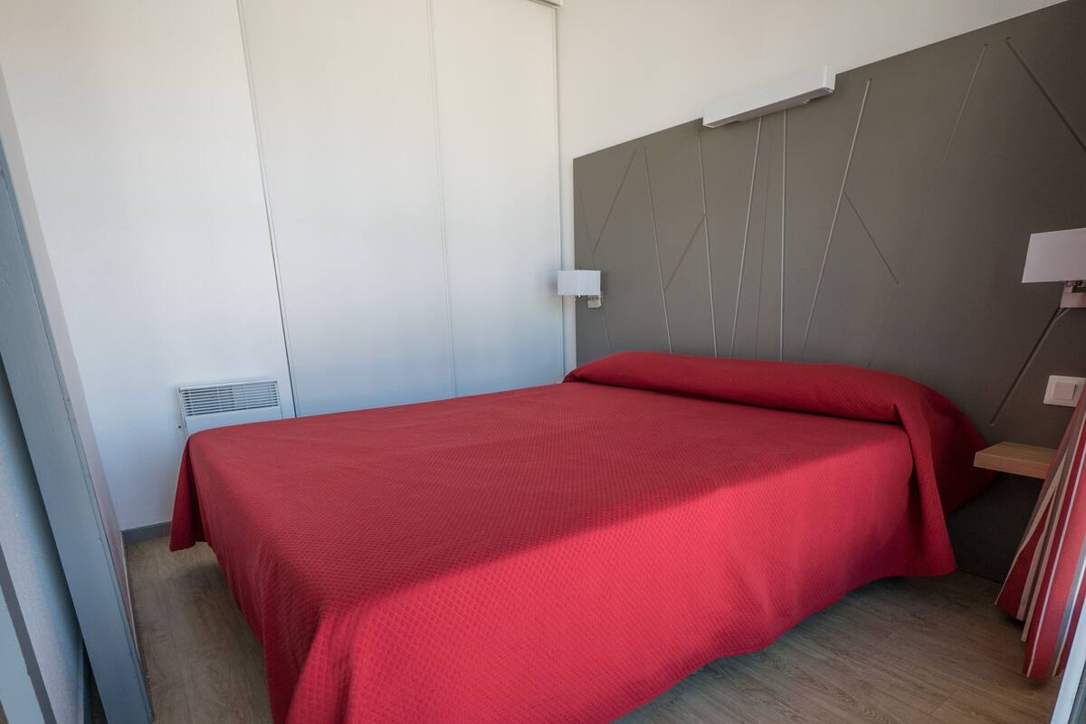 A single bed with a red bedspread in a room with a grey headboard and a white wall.