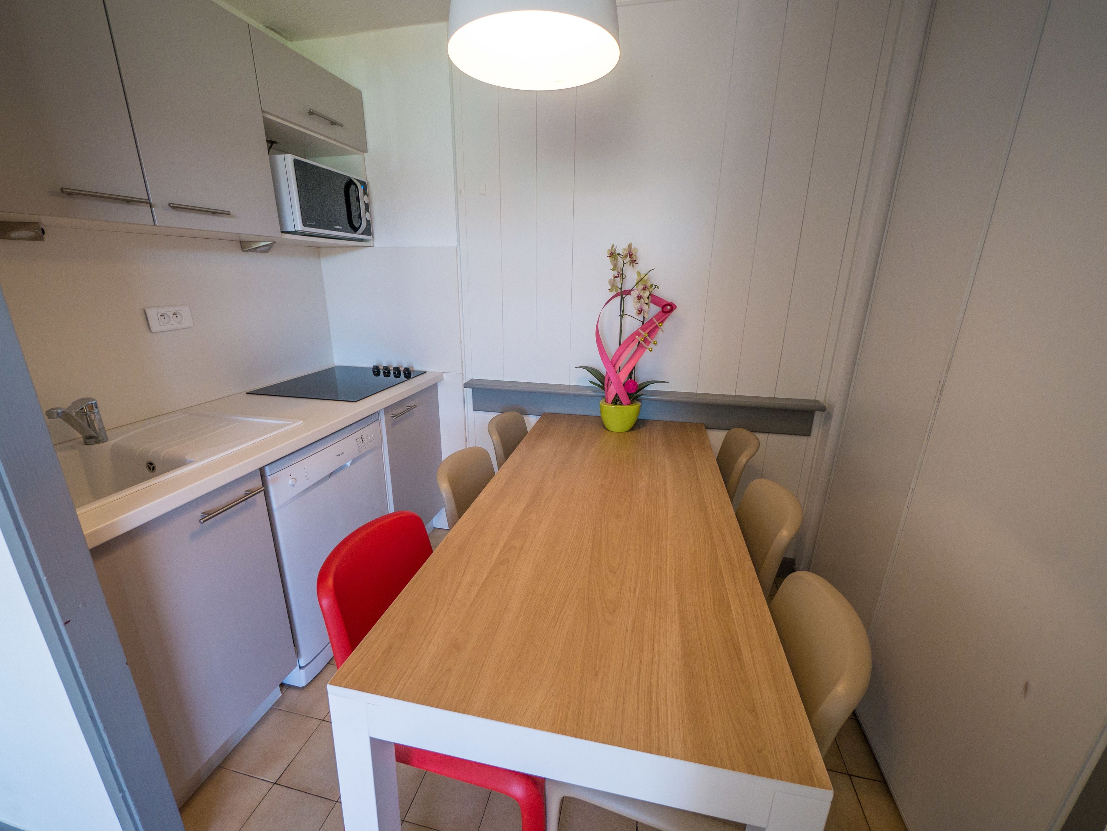 Una cocina compacta con microondas, lavamanos y lavavajillas pequeño. Hay una mesa de comedor de madera con cuatro sillas y un florero con flores.