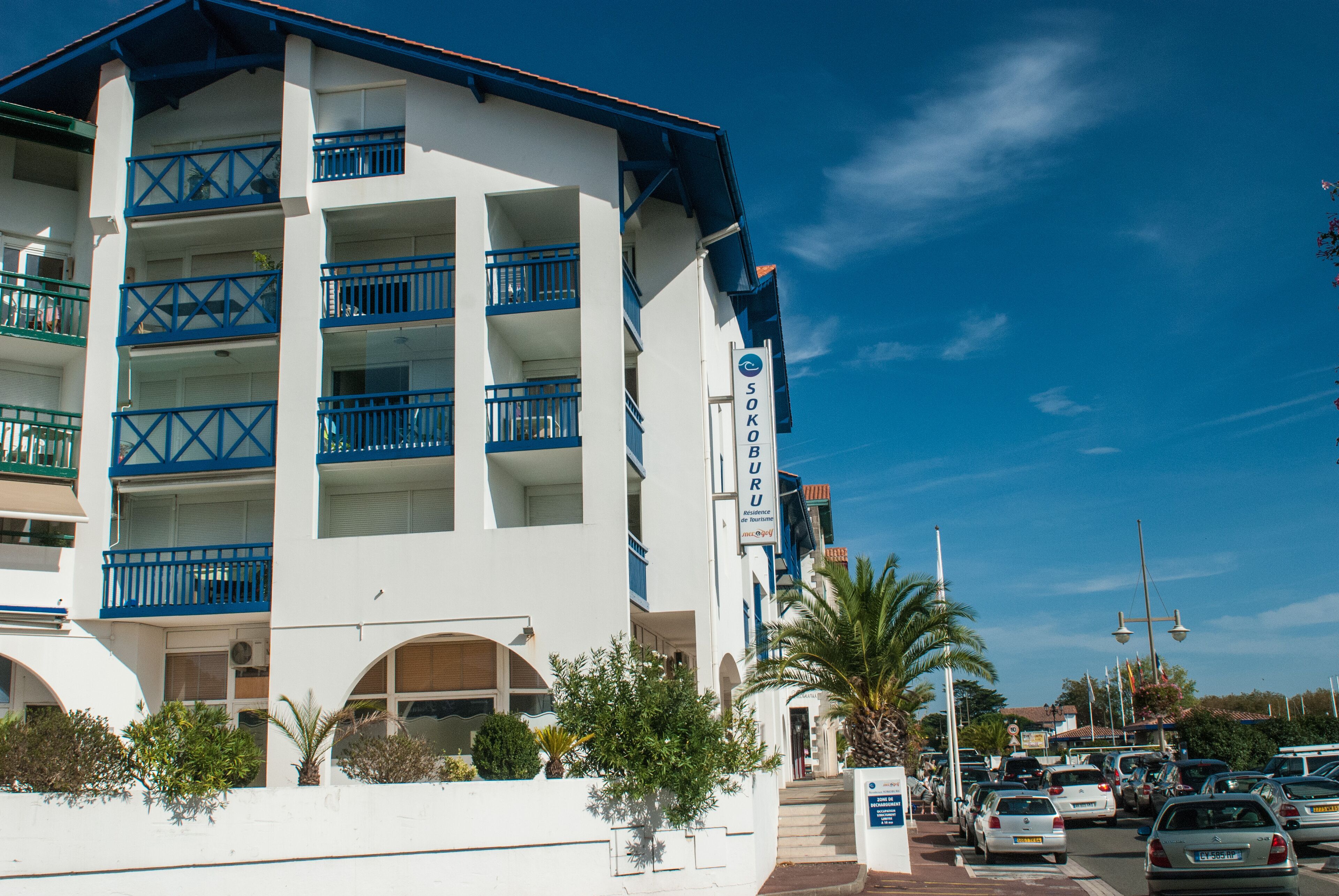 Un hotel de varios pisos con balcones, un cartel que dice "SOKO BURU" y un estacionamiento con coches.