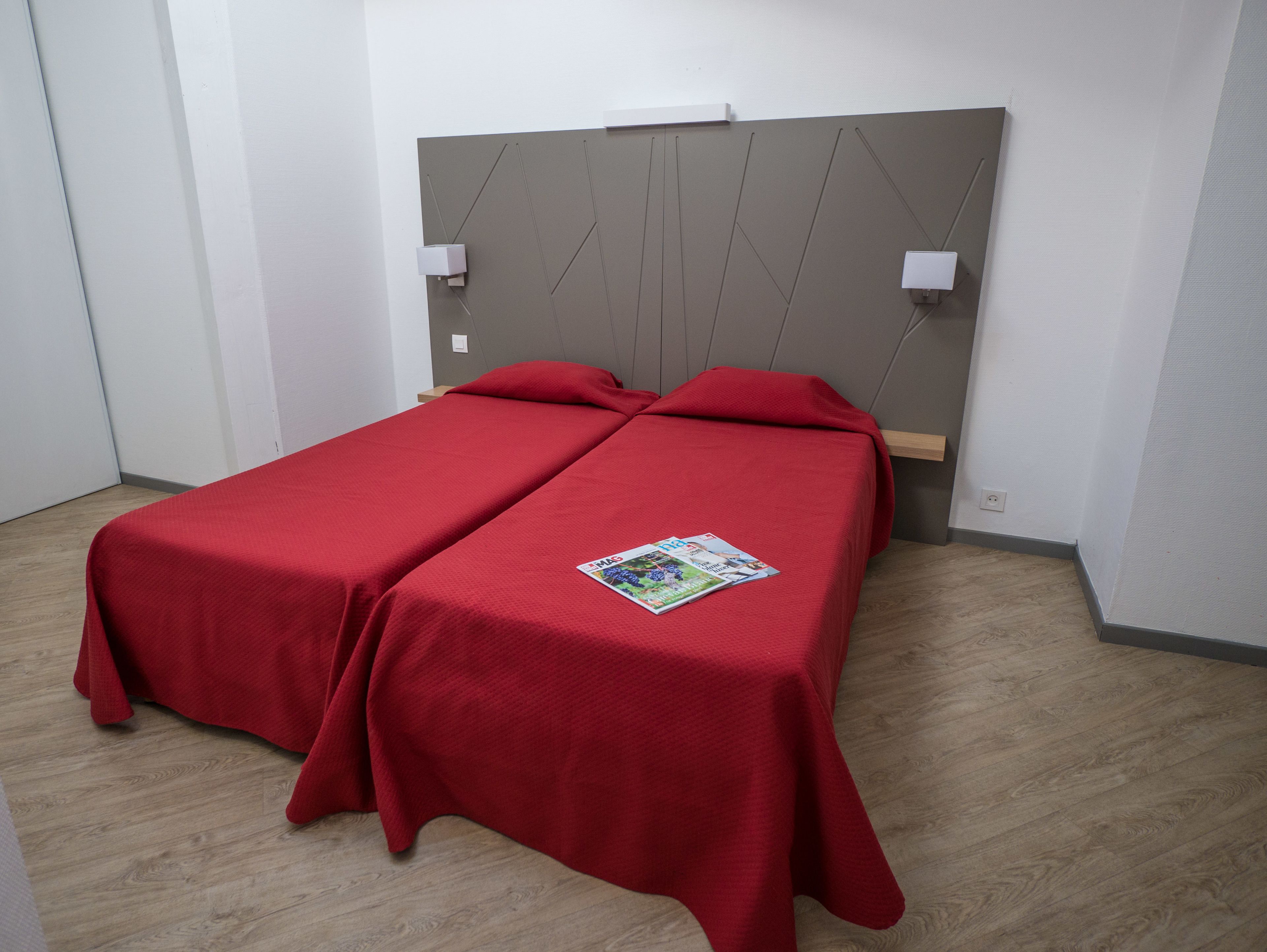 A room with two single beds, each with a red bedspread and a magazine on one bed. There are two bedside lamps and a wooden headboard.