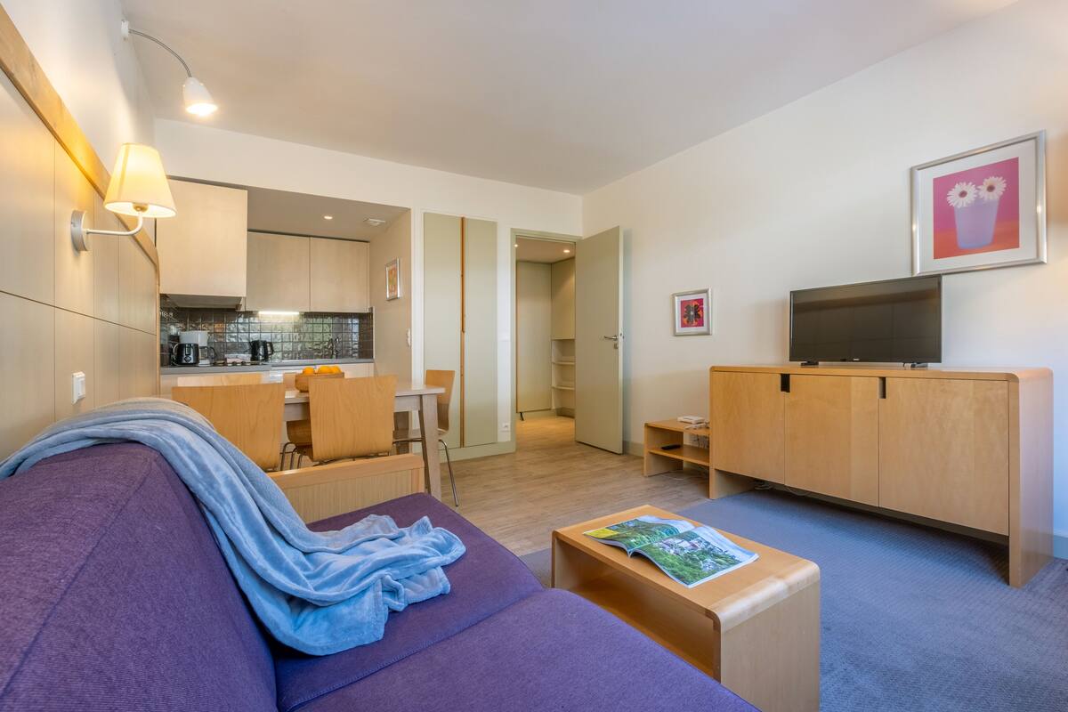 Ein modernes Wohnzimmer mit einem violetten Sofa, einem Holz-TV-Schrank und einer Küche im Hintergrund.