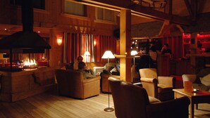 Een knus, gedimd verlichte loungeruimte met houten wanden en een open haard. Er staan meerdere leren fauteuils en een bank, en een bar met een persoon achter de toog.