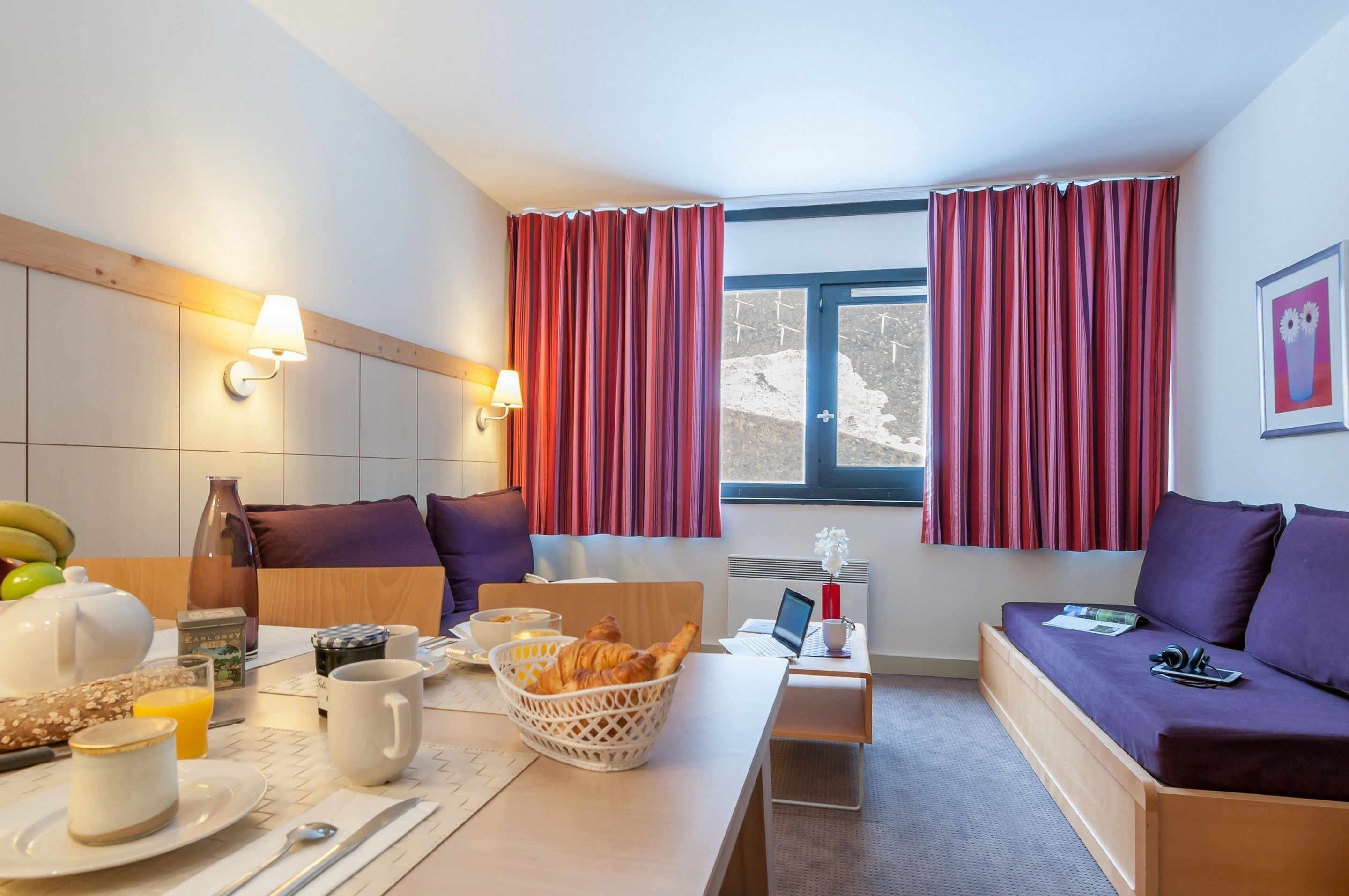 A hotel room with a breakfast setup, a sofa, a laptop, and a view of the outdoors.