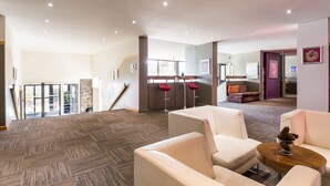 Een ruime lobby met een tapijtvloer, een zithoek met witte banken, een bijzettafeltje en een klein decoratief object. Er staan twee rode barstoelen en een stenen zuil.