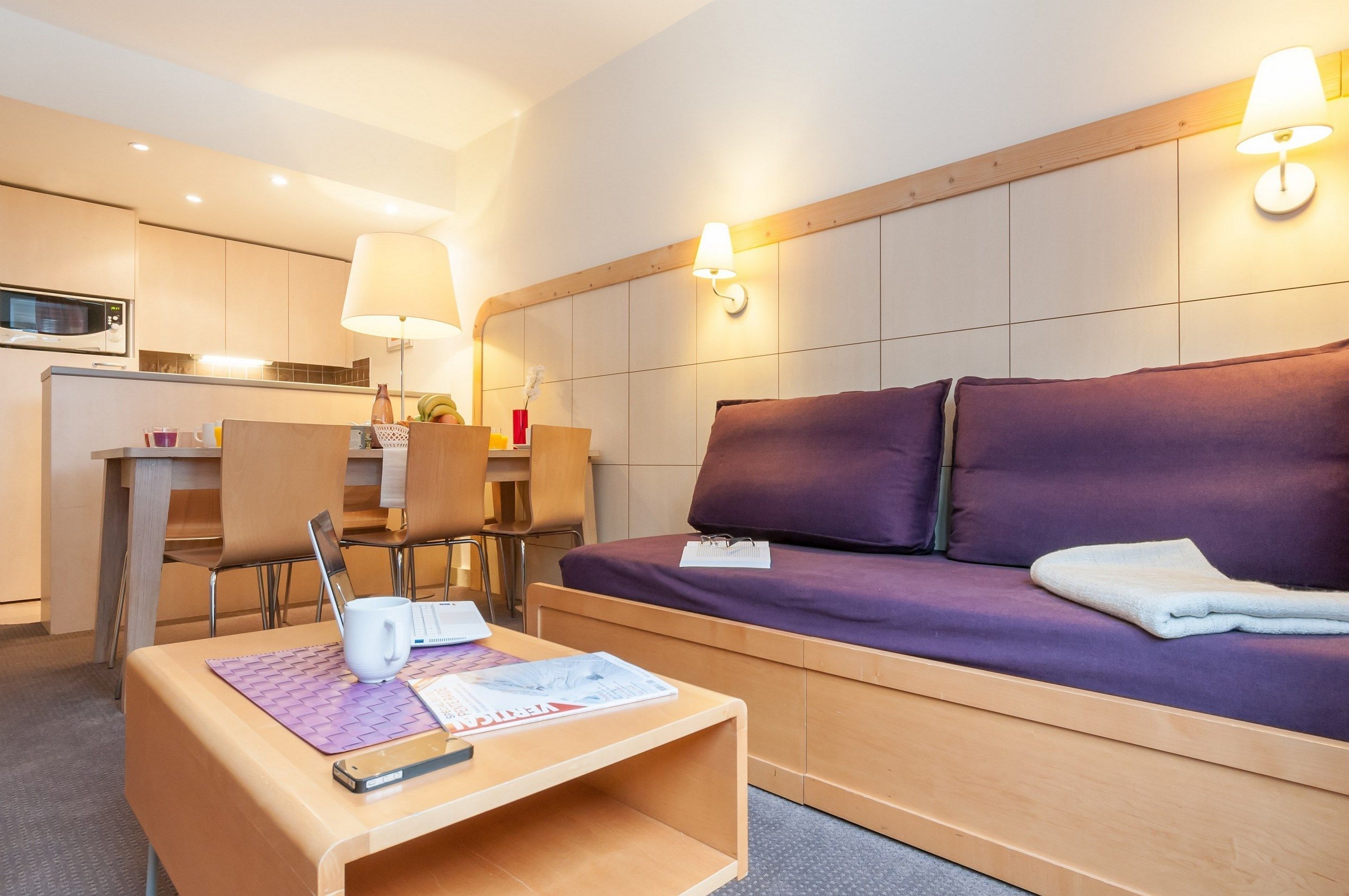 Ein modernes Hotelzimmer mit einem violetten Sofa, einem Holztisch mit einem Laptop und einer Tasse, einem Schreibtisch mit Stuhl und einer Mikrowelle im Küchenbereich.