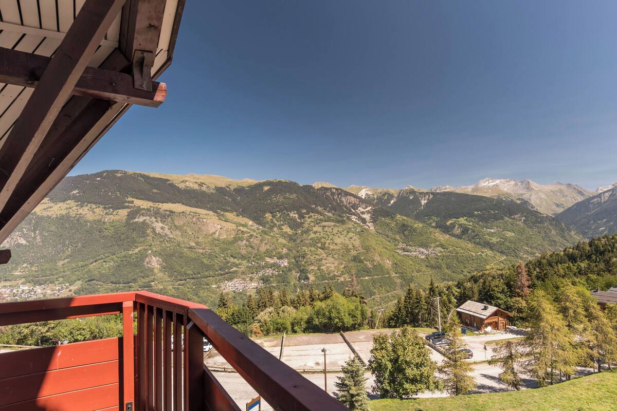 Blick auf Berge vom Balkon mit roter Brüstung, ein kleines Gebäude und ein Parkplatz.