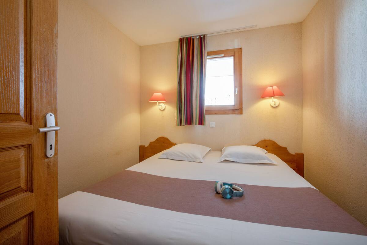 Une chambre d’hôtel avec une porte en bois, un lit avec une literie blanche et marron, deux lampes de chevet et une fenêtre avec un rideau.